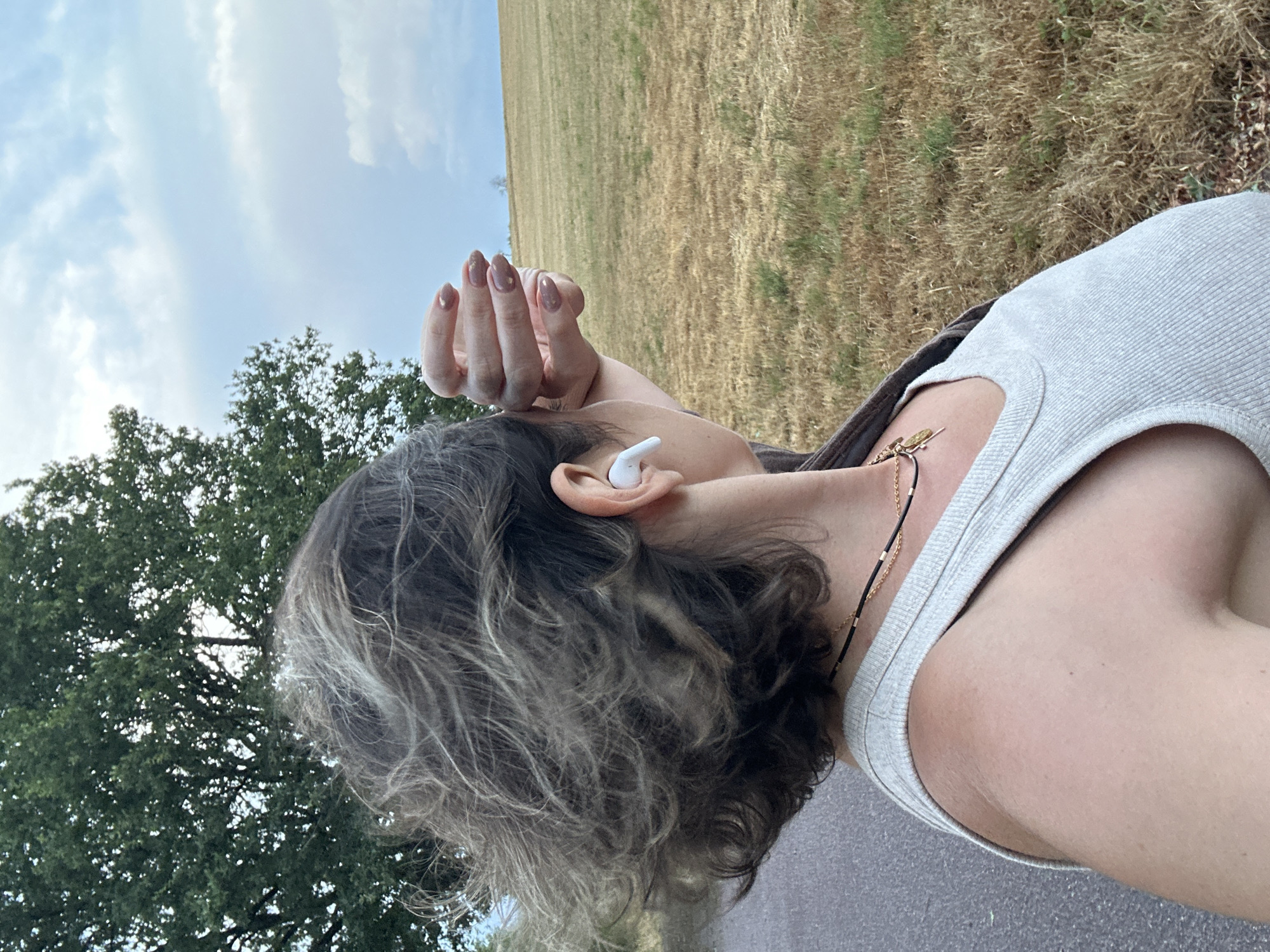 A person, viewed from the right side, stands on an asphalt path in Wadern, Germany. The person has short, tousled brown and gray hair, and is wearing a light gray ribbed tank top and a dark jacket draped over their shoulders. They have white wireless earbuds in their right ear and are touching their forehead with their left hand, revealing fingernails painted a brown color with small gold accents. In the background, a large, green tree stands to the left, and a dry, grassy field stretches to the right under a cloudy sky.