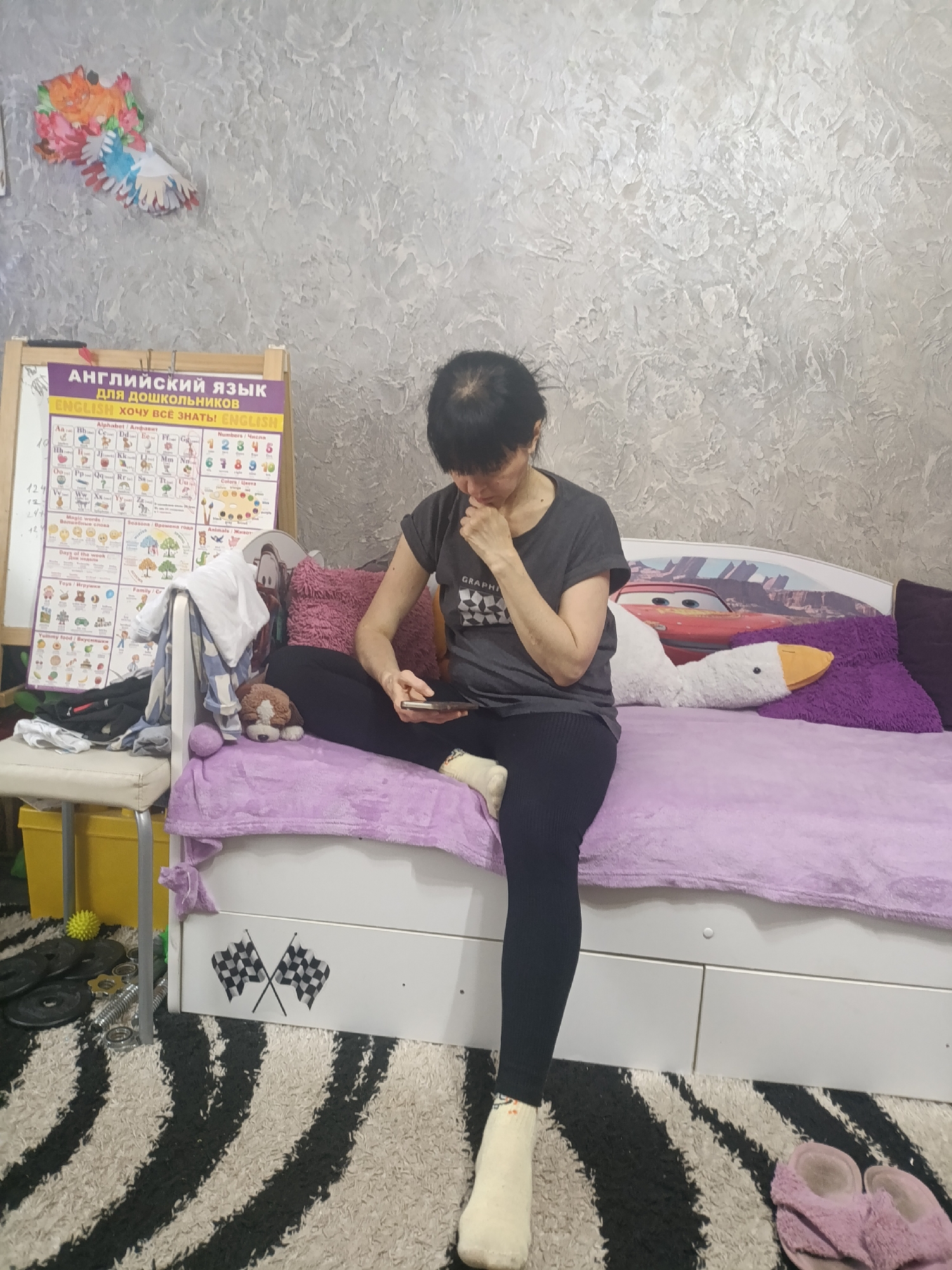 A middle-aged woman with short, dark hair is sitting on the edge of a bed in a room. She is wearing a gray t-shirt and black leggings. Her right leg is bent at the knee with her foot planted on the floor, clad in a light-colored sock. She holds a smartphone in her left hand, looking down at the screen while her right hand is raised to her mouth, appearing thoughtful or concerned.
The bed is covered in a light purple blanket, with purple and pink pillows scattered behind her. A stuffed white goose is positioned next to her. The headboard of the bed features an image of a red race car.
To the left of the woman is a small white chair with a yellow storage container underneath. Various items are draped over the chair, including clothes and what appears to be a stuffed animal. Behind the chair is a whiteboard displaying a colorful English language learning chart with Russian text.
The room has textured, gray-toned walls. The floor is covered in a black and white zebra-striped rug. A pair of pink slippers lie on the rug near the foot of the bed. The bottom of the bed frame features drawers decorated with checkered flag patterns.
Based on the whiteboard containing Russian and English text, the location is likely in a Russian-speaking area. While the exact location within Krasnodar, Russia, is not identifiable from the image, the room appears to be in a residential setting, possibly a child's bedroom.