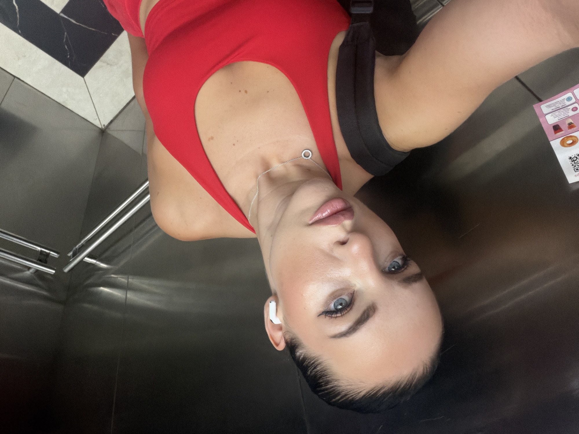 A selfie captures a woman from an overhead perspective inside an elevator, appearing to be rotated 90 degrees counter-clockwise. The woman, who has blue eyes, dark eyebrows, and light-colored lipstick, is looking directly at the camera with a neutral expression. Her dark hair is pulled back and slicked down around her temples. She is wearing a red top or bodysuit, and a silver necklace with a small circular pendant hangs around her neck. A white wireless earbud is visible in her right ear, and a black backpack strap is visible over her left shoulder.
The elevator's interior features polished metallic walls, reflecting some light. The floor, visible in the upper left corner, has a distinctive black and white geometric tile pattern. Metallic handrails are also visible on the elevator wall. In the background, to the right of the woman's head, a small, partially obscured pink and white flyer or sign is affixed to the wall. The word "DONUT" is clearly legible on this flyer, along with unreadable smaller text and an illegible QR code. The setting is consistent with an urban environment, specifically in Bac Nha Trang Ward, Vietnam.