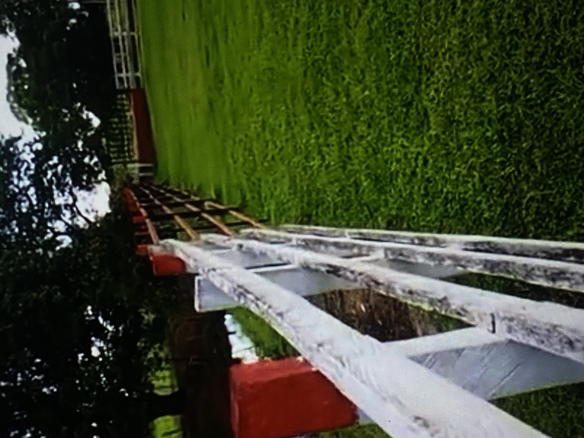 A wide shot captures a section of a white wooden fence bordering a lush green grassy field. The fence, with a weathered appearance, is in the foreground, running diagonally across the frame. To the left of the fence, a red rectangular marker is visible. In the background, mature trees with dark foliage create a dense canopy. Further in the distance, another white fence is partially visible. The scene appears to be an outdoor rural or park-like setting, likely located in the city of Tinaquillo, Venezuela. No people or animals are discernable in the image, and no actions or interactions are depicted. The overall lighting suggests an overcast or twilight condition.