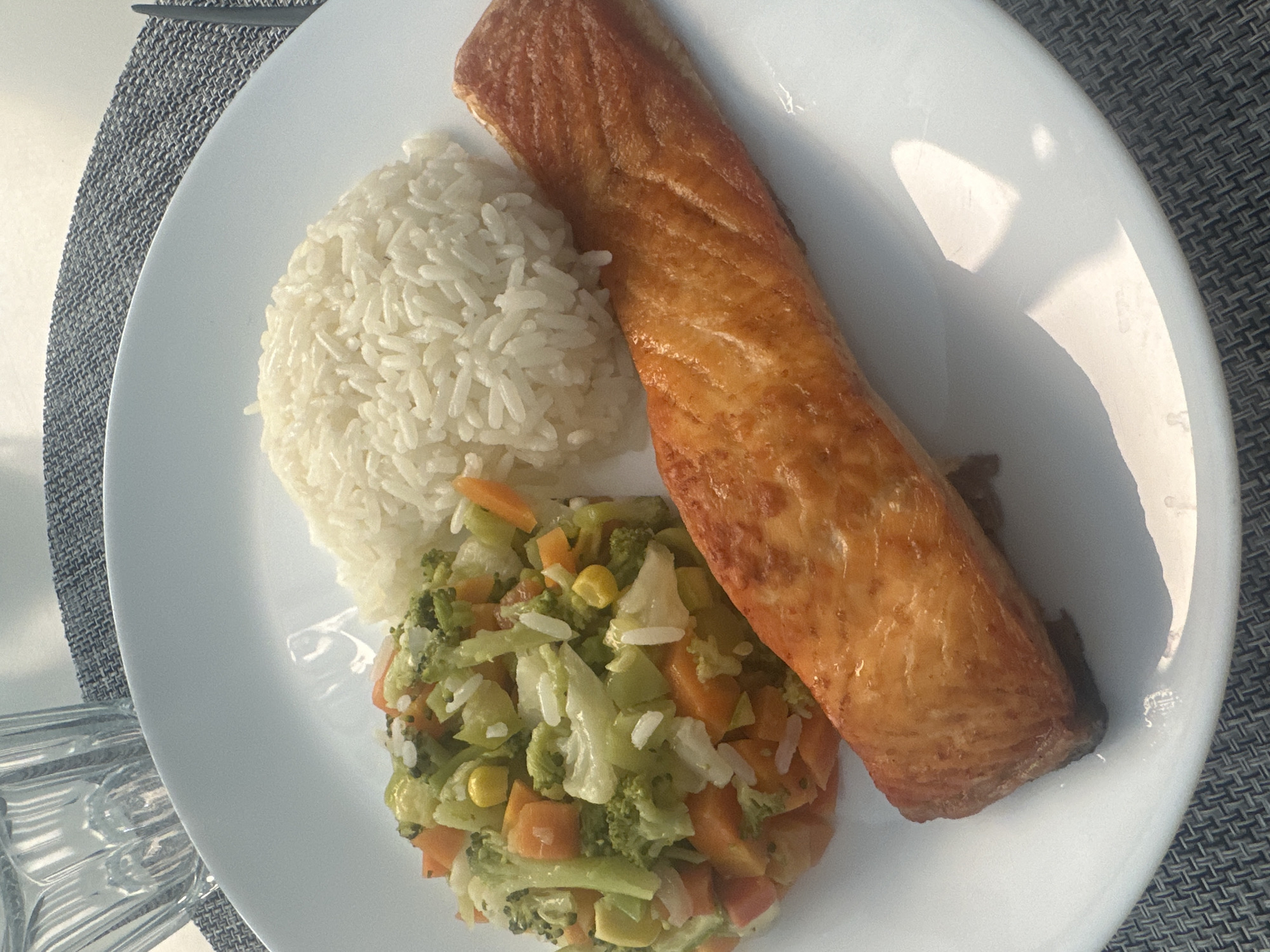 A close-up, top-down view captures a prepared meal served on a white, oval-shaped plate. The plate holds a large, golden-brown piece of cooked fish, likely a salmon fillet, which appears to be pan-fried or baked. Adjacent to the fish is a neat mound of fluffy white rice. Below the rice and next to the fish is a serving of mixed vegetables, including visibly chopped orange carrots, green broccoli florets, white cauliflower pieces, and yellow corn kernels. Some loose grains of white rice are scattered over the vegetables.
The plate rests on a gray, woven placemat, which has a distinct checkered or basket-weave texture. A clear glass, partially visible on the far left, sits on the same placemat. The scene is brightly lit by strong natural light, casting distinct highlights and reflections on the shiny surface of the plate and subtle shadows, indicating it is daytime. No people or visible text are present in the image. The setting suggests a casual dining experience, possibly at a table in Puebla, Mexico.