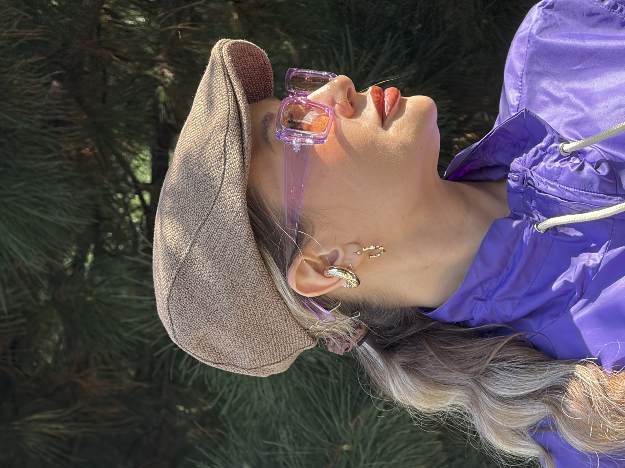 A woman with fair skin, wearing a brown newsboy cap and purple sunglasses with clear frames, is turned upwards towards the sun. She has on bright red lipstick, a nose ring, and gold earrings. She wears a purple nylon jacket. The background is a blur of green foliage, possibly trees, indicating an outdoor setting. No location information is discernible.