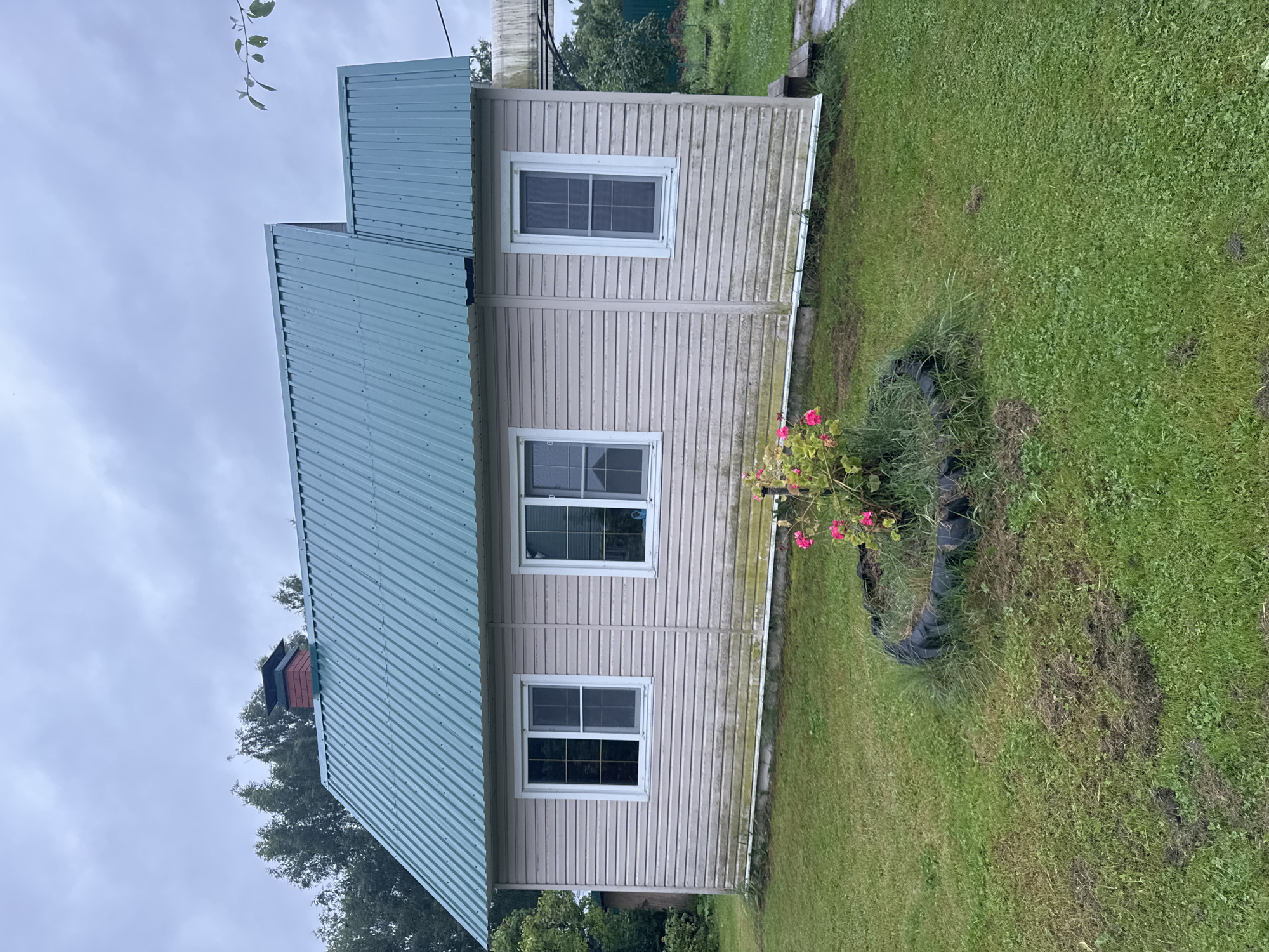 The image shows a modest, single-story house with a teal-colored metal roof and light-colored vinyl siding. The house has several windows with white frames. A red-bricked chimney with a black cap is visible on the left side of the roof. In front of the house, a small circular flower bed is outlined by a repurposed tire and contains pink flowers and green foliage. The surrounding area is a grassy yard, with some patches of less manicured grass and what appear to be molehills. The sky is overcast and cloudy. The house is located in the city of Krasnoye, Russia.