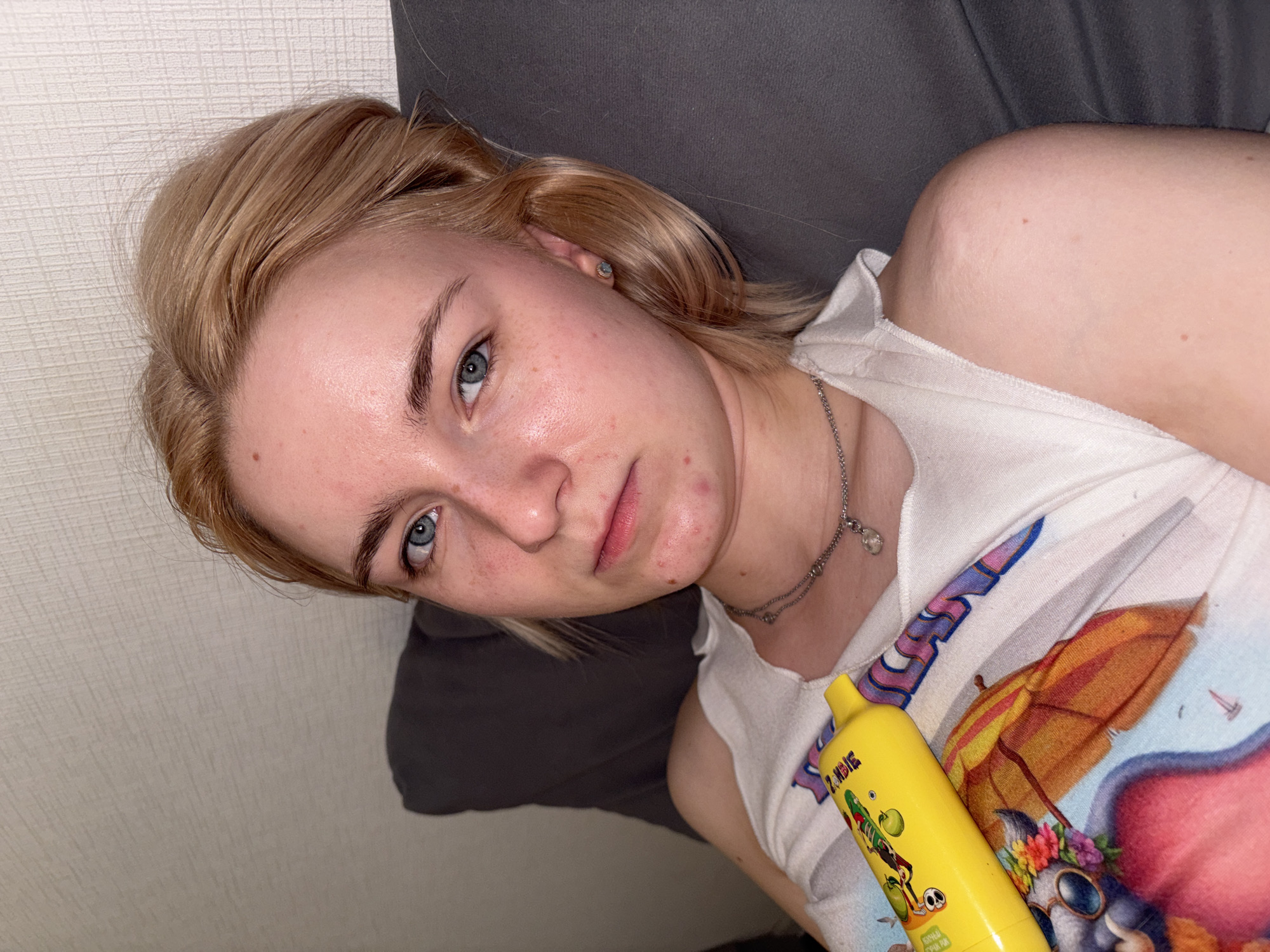 A young adult female, appearing to be in her late teens or early twenties, is depicted reclining on a dark grey cushion against a light-colored, textured wallpaper in a residence in the city of Unknown, Russia. She has blonde hair, blue eyes, and fair skin with visible blemishes and freckles across her face. Her expression is neutral, with a slight downturn to her mouth. She wears a white sleeveless top featuring a multi-colored graphic print of a cartoon character with a floral headband and sunglasses under an umbrella. Two layered silver-colored necklaces, one with a small clear stone pendant, are visible around her neck. A small stud earring is present in her right ear (viewer's left). In her right hand (viewer's left), she holds a bright yellow cylindrical object, identified as a disposable vape device, with the brand name "ZOMBIE" and Cyrillic text visible on its label. The lighting suggests an indoor setting.