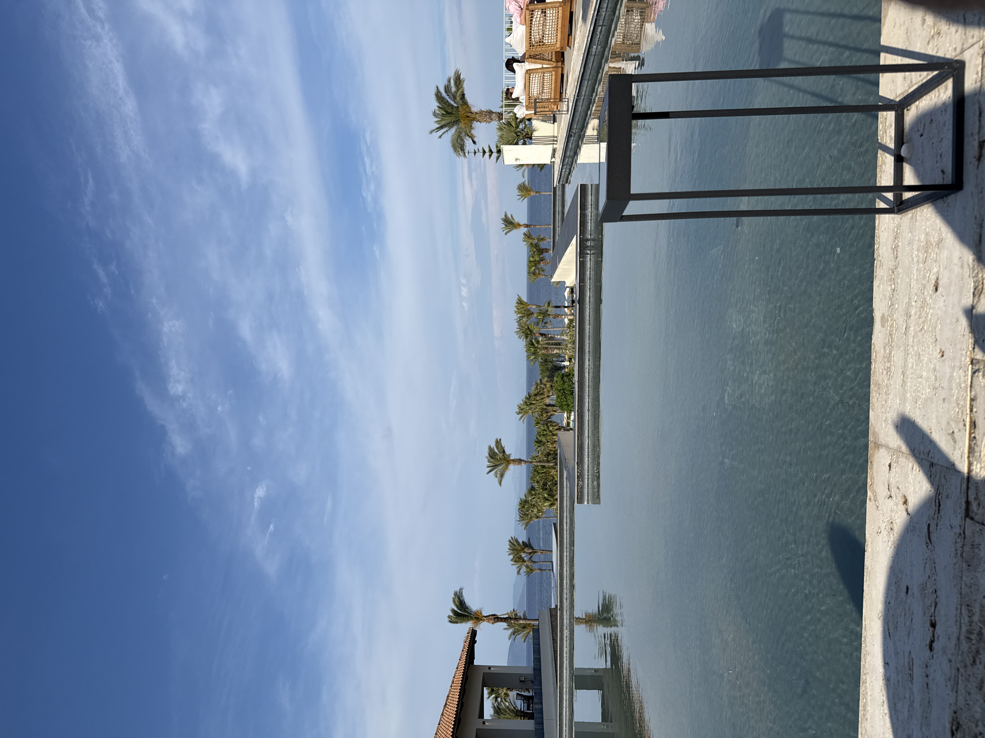 The image depicts a coastal scene, possibly a resort or upscale area, in Kos, Greece. The horizon features a visible coastline, and a series of palm trees extends into the distance. These trees are reflected in the water, indicating a tranquil water surface. The sky is blue with some white clouds, suggesting fair weather.
There are some people sitting and relaxing at the resort.
A structural element, a black rectangular frame, appears in the foreground. The scene is shot from a higher vantage point, possibly from an elevated balcony or building. The overall setting conveys a sense of luxury and relaxation.