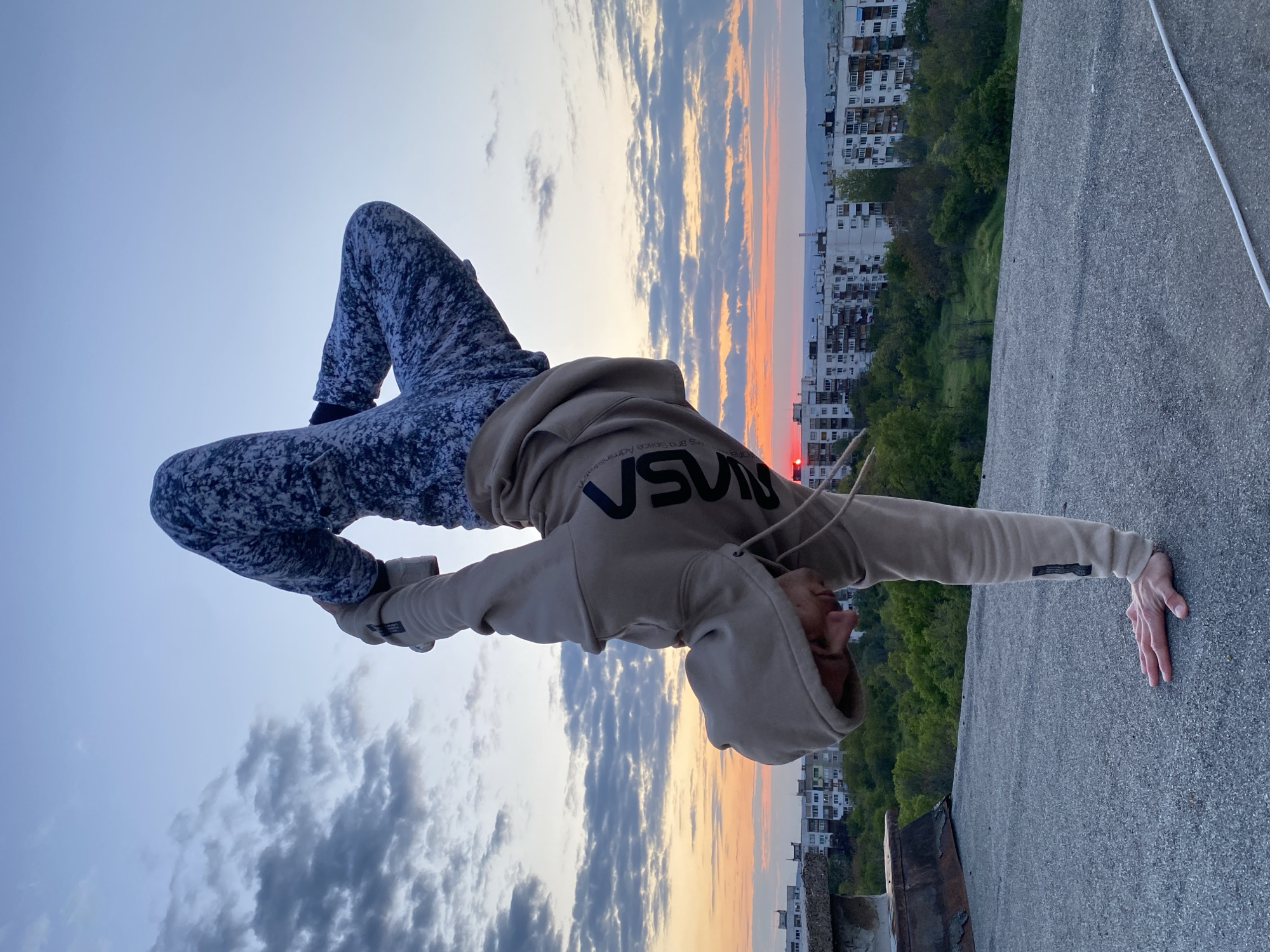 A person is doing a handstand on the edge of a building, likely in Pleven, Bulgaria. The person is wearing a hooded sweatshirt with the letters "NASA" printed on it, and patterned pants. They are leaning forward, with one hand on the edge of the building. The background consists of a sunset sky with clouds and a cityscape. Buildings and trees are visible in the distance. The concrete surface of the rooftop is visible.