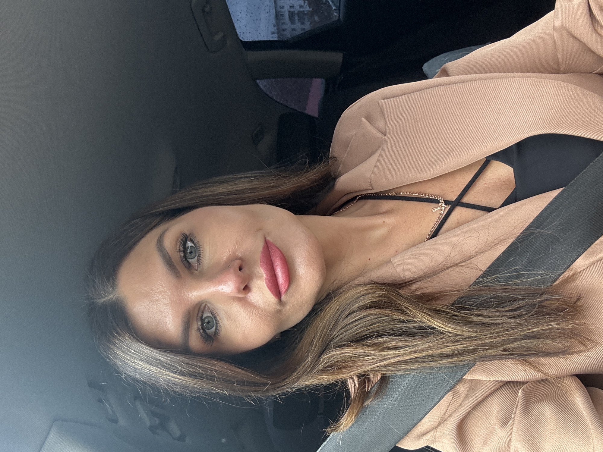 A woman is captured in a selfie-style shot, seated in what appears to be the passenger seat of a car, based on the visible seatbelt and interior. The image is oriented with the woman's head tilted to her right.
The primary subject is a woman with long, flowing brown hair, styled with some lighter highlights. She has striking green eyes, dark eyebrows, and is wearing pink lipstick. Her makeup appears neat and natural. She is wearing a black top with delicate criss-cross straps visible at the neckline, beneath a light beige or pale peach-colored blazer or jacket. A thin gold necklace with a small, cross-like pendant hangs around her neck. Her expression is calm and direct, looking towards the camera.
The car's interior is visible, featuring a dark gray or black headliner and a dark seatbelt diagonally across her chest and shoulder. Through the car window in the upper left background, hints of an urban environment, possibly a building, are discernible. The window itself shows streaks or droplets, suggesting it might be raining or has recently rained. This detail, combined with the subdued natural lighting, indicates an overcast day, potentially in Chaikovsky, Russia, as per the provided context. There is no visible text in the image.
