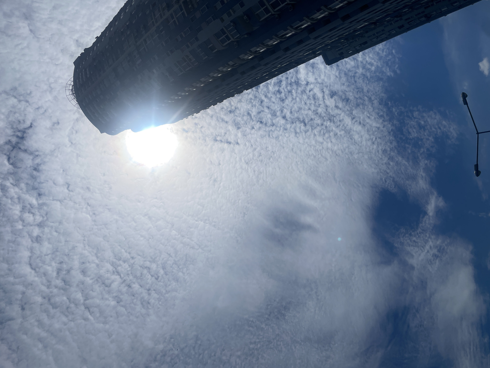 An upward shot of a dark, tall building against a blue sky with scattered white clouds. The sun is prominently featured, partially obscured by the building and emitting a strong glare. The building appears to be residential or office, with visible windows and air conditioning units. A portion of another building is visible on the right side. In the lower portion of the frame, a street lamp with two fixtures is visible. The image was taken in Kyiv, Ukraine.