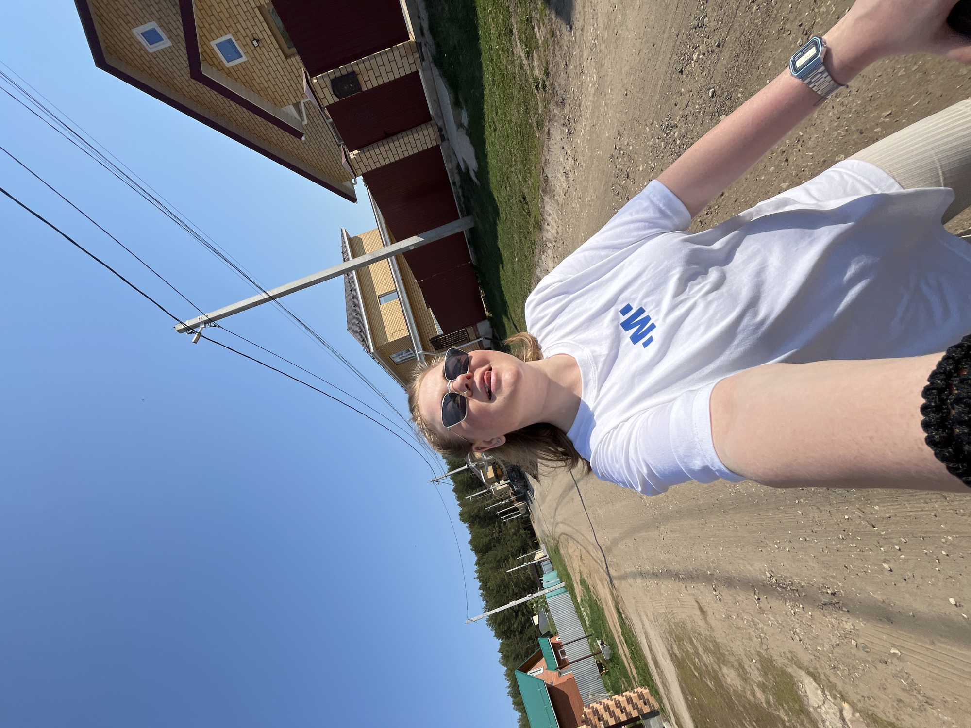 A person is standing on a dirt road in Chaikovsky, Russia. They are wearing sunglasses and a white t-shirt with a blue logo. Their right arm is visible, and they are wearing a watch on their wrist. Behind them, there are houses with brick and metal siding and green roofs, and a line of trees. Power lines run above the road. The sky is bright blue.
