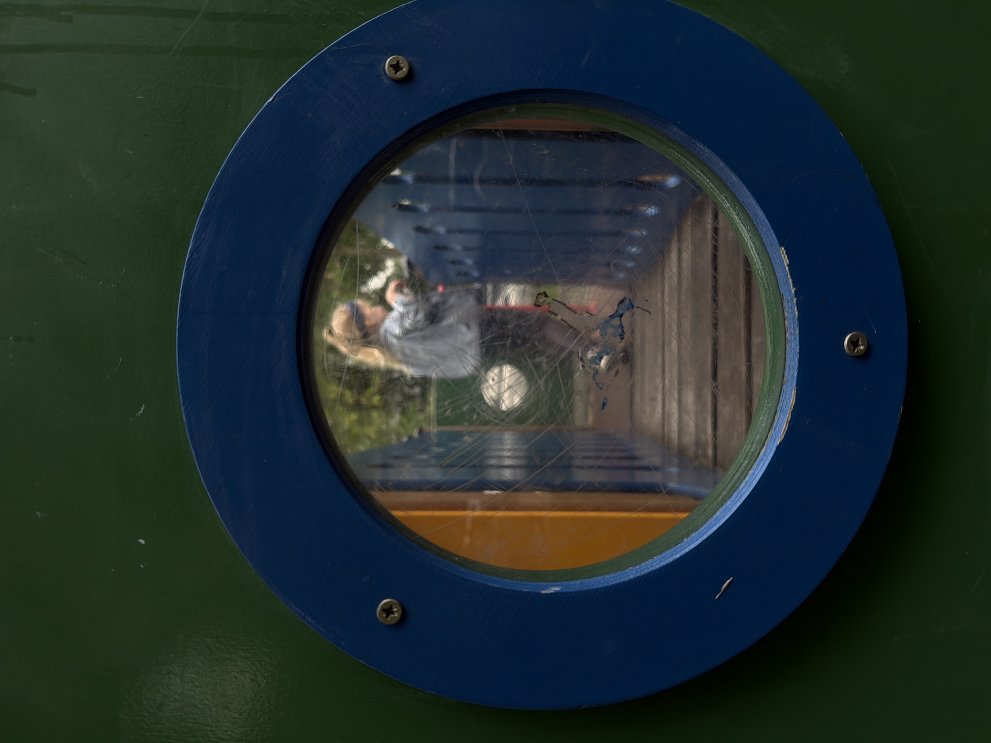 The image shows a close-up of a circular window with a blue frame, set into a dark green surface, likely part of a playground structure. Through the scratched plastic window, a reflection is visible. This reflection shows a person, possibly a child, wearing a light-colored shirt. The person appears to be looking into the window. In the background of the reflection, blue and yellow surfaces are visible, suggesting the interior of a playground or playhouse. The scene is outdoors, with some greenery visible beyond the structure, indicating daytime. The scratches on the window obscure some details of the reflection. The overall impression is a playful and slightly distorted view of a moment at a playground. There is no visible text in the image. The context of Moscow, Russia is provided but not visually evident in the image itself.