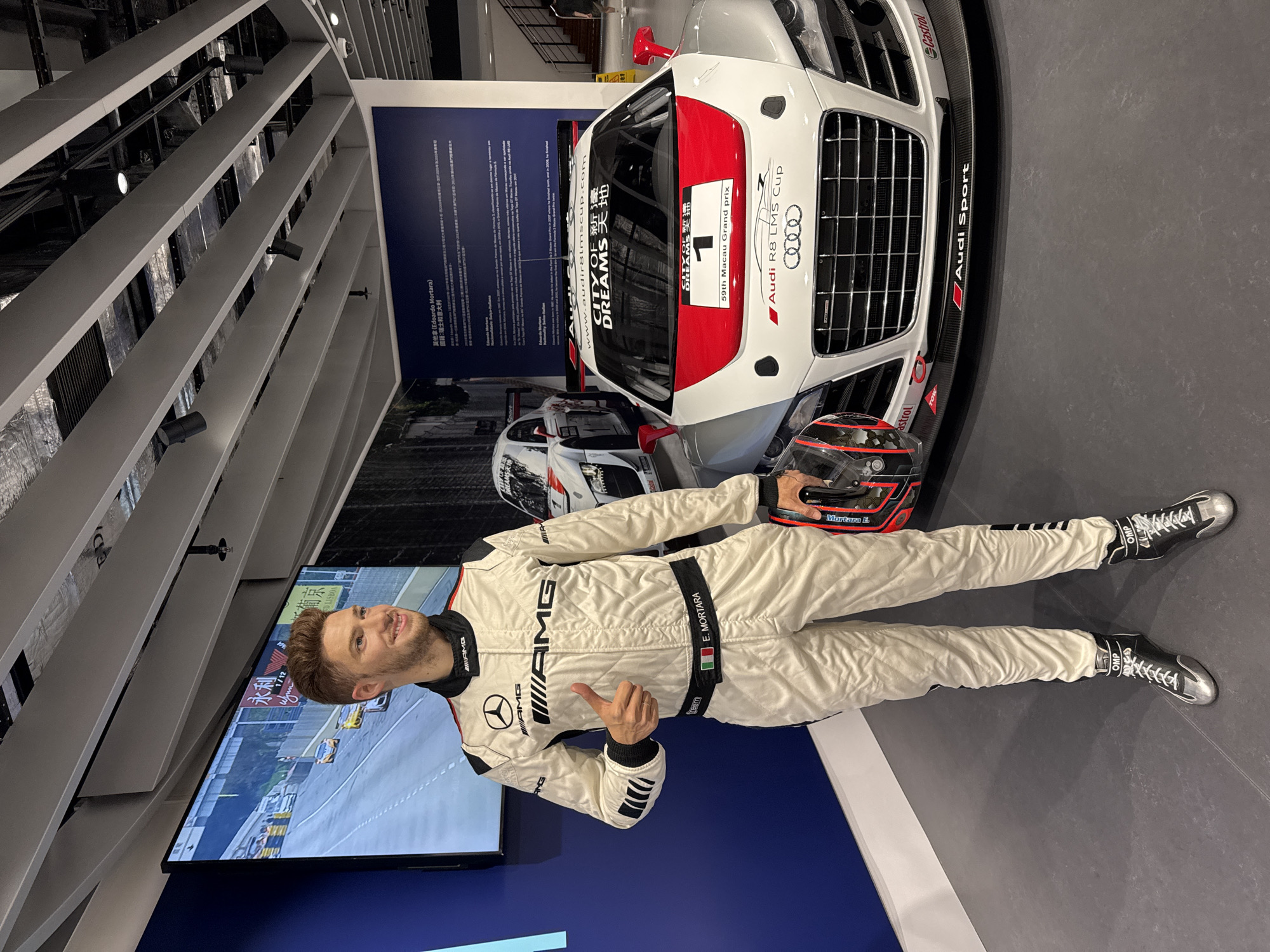 Here is a detailed description of the image:
The image captures a scene inside what appears to be a car exhibit, possibly in Macau, China. A man, dressed in a white AMG racing suit and black and silver racing shoes, is the primary subject. He's giving a thumbs-up gesture. Next to him, on the right, is a red and black racing helmet.
Behind the man is a silver and red race car, an Audi R8 LMS Cup, marked with the number "1" and the text "59th Macau Grand Prix". "Audi Sport" is labeled on the car's front. The backdrop features a display about the driver and his car, including information about Edoardo Mortara (other拿(Edoardo Mortara)國籍:瑞士和意大利). There's also a TV screen on the left, showing what seems to be a racing game or simulation, with the time "1/12" visible on the screen.
The environment includes architectural details like beams and a modern interior design. The location may be somewhere with an association to the "City of Dreams" as the car's advertisement points out: www.audir8lmscup.com CITY OF DREAMS孺