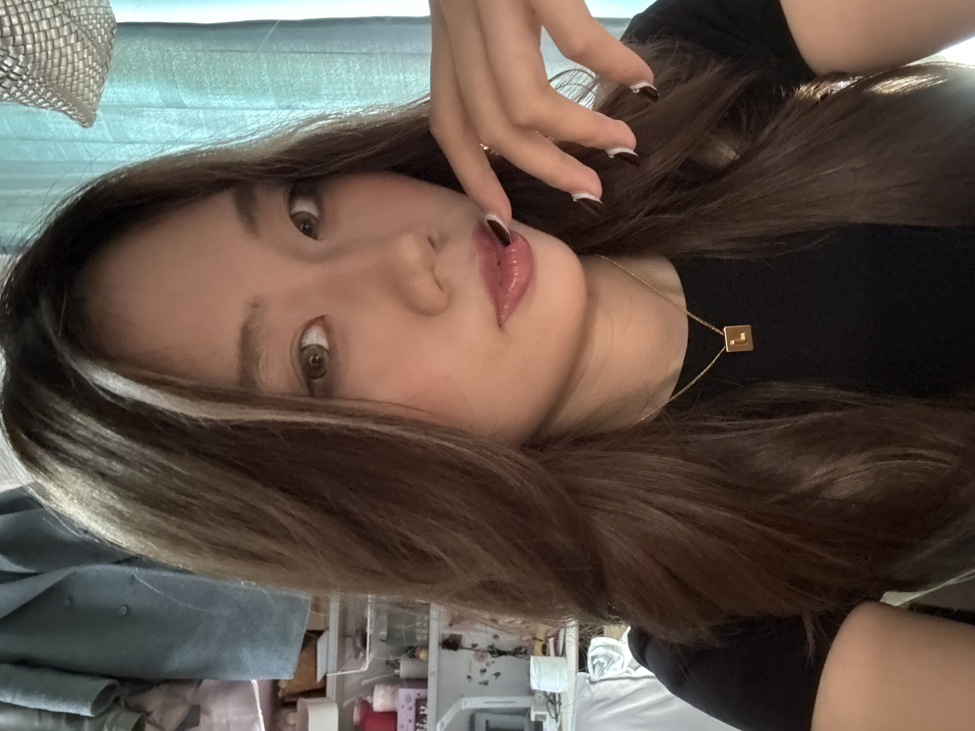 A selfie of a young woman with long brown hair with blonde streaks. She is wearing a black shirt and a gold necklace with a square pendant. Her left hand is raised with a finger touching her lips, which are glossy with lip gloss. Her nails are painted with a black and white design. The background is a cluttered interior space with a white wall and shelves filled with various items. The location is indicated to be Nan'an District, China.