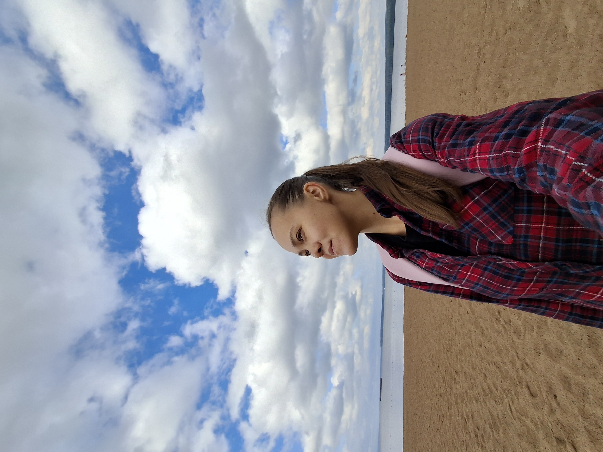 A young woman with a ponytail, wearing a red and blue plaid shirt and a pink backpack, stands on a sandy beach. She is looking to her left with a slight smile. The background shows a body of water with a distant shore and a partly cloudy sky. This scene appears to be the Kama River embankment in Chaikovsky, Russia, a popular recreational area.