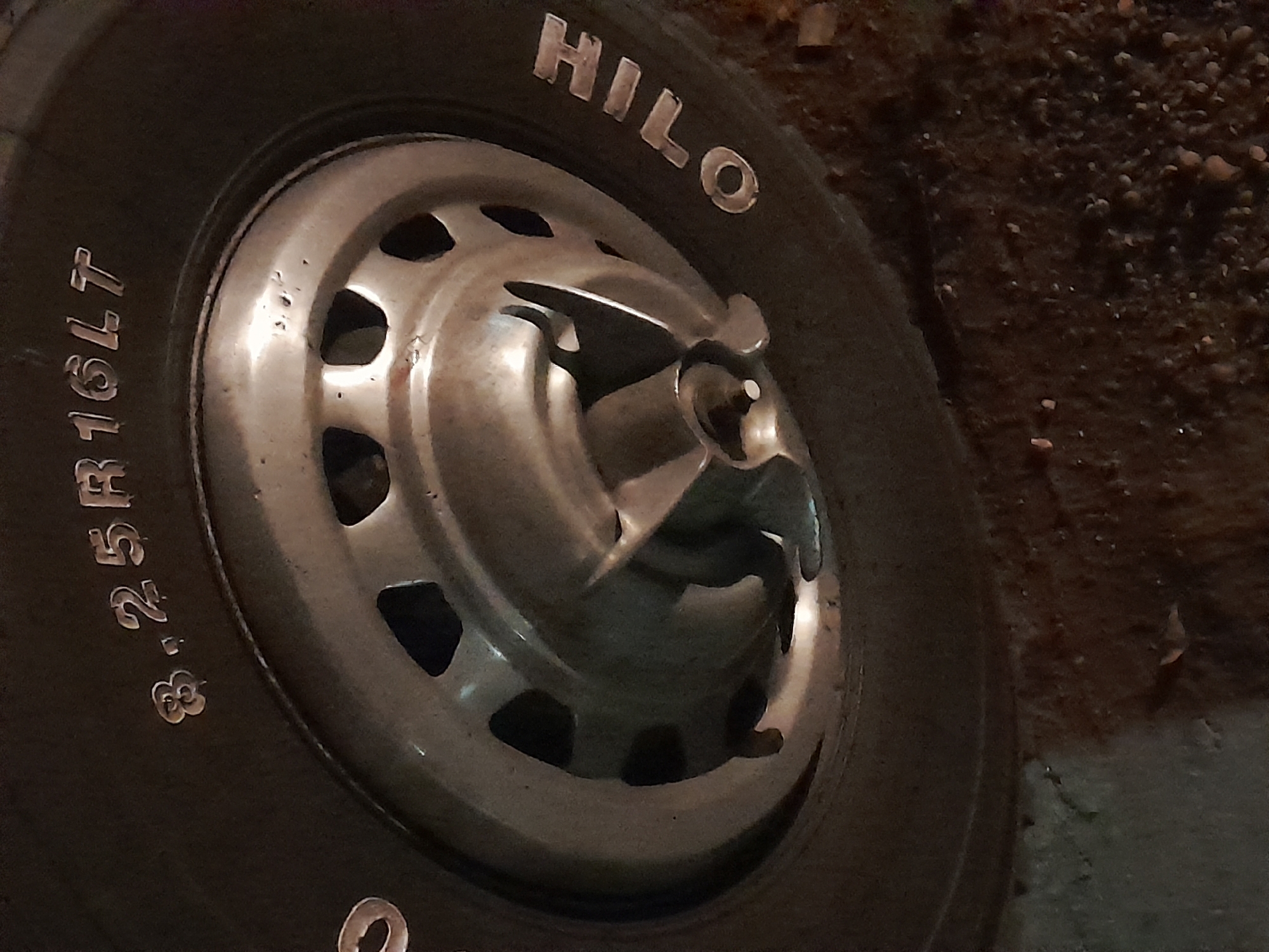 The image shows a close-up of a tire on a vehicle, specifically the brand "HILO" which is written in white lettering on the sidewall. The tire size is also visible, reading "8.25R16LT". The tire is mounted on a silver wheel with visible lug nut holes and a hubcap. The background is dark and appears to be asphalt or dirt, suggesting the vehicle is parked outdoors. There are no people visible in the image. The location of Santa Ana, Venezuela is not depicted or suggested by any elements in the photograph.