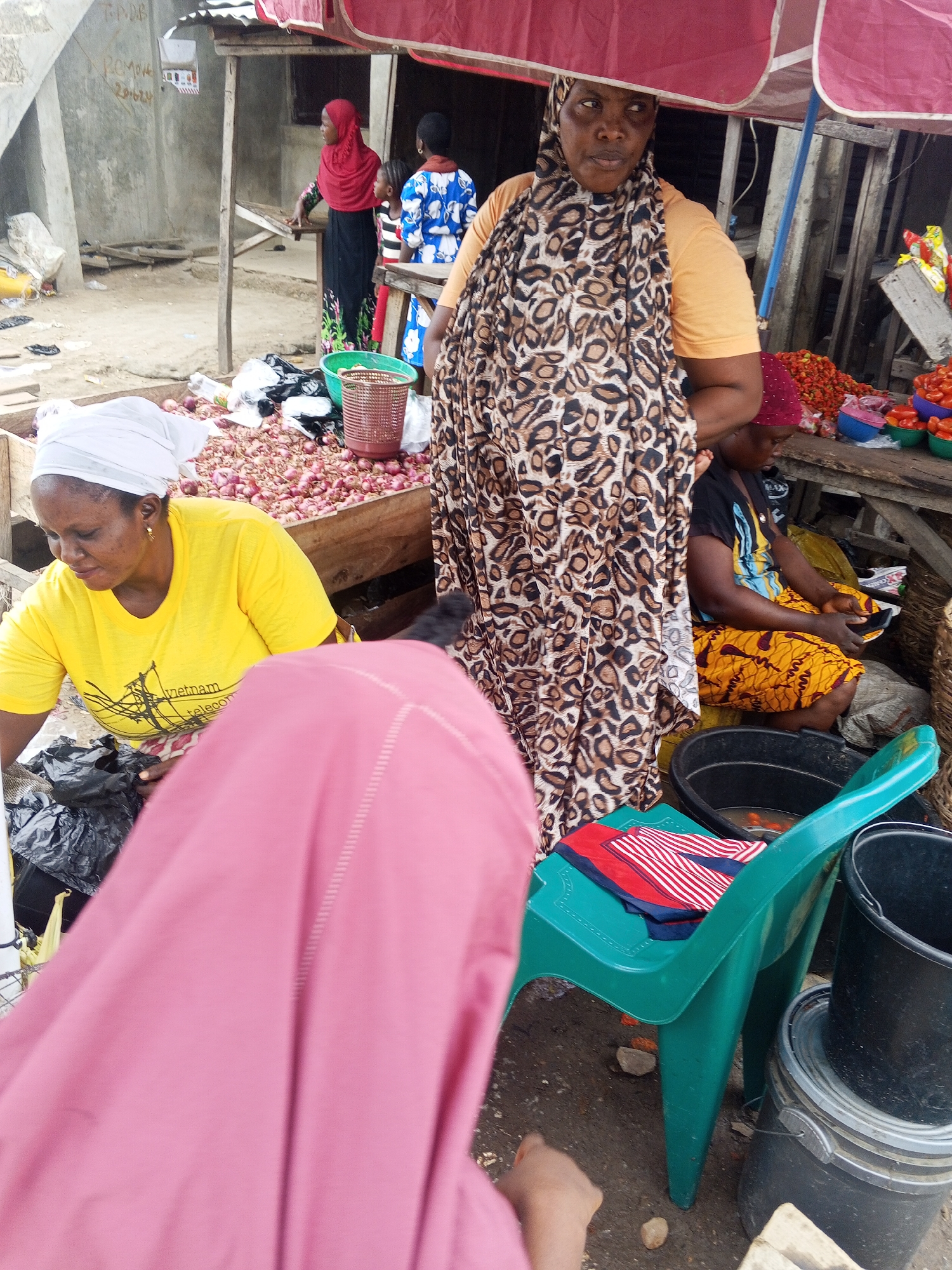 The image depicts an outdoor market scene in Usunwe, Nigeria. In the foreground, a person wearing a pink hijab is partially visible. To their right, a woman in a yellow t-shirt and white headscarf is looking down. Behind her, a woman in an orange shirt and a patterned leopard print hijab stands, looking towards the left. To her left, a child in a blue and white patterned shirt is visible. Further back, other individuals are present, some wearing hijabs and traditional Nigerian clothing.
The market stall features various goods, including piles of onions and tomatoes displayed in wooden crates and plastic bowls. A red umbrella provides shade over the stall. Several plastic buckets, one green chair with folded fabric on it, and baskets are also present. The ground appears to be dirt and gravel. The background shows a concrete building and wooden structures, indicative of a market setting.