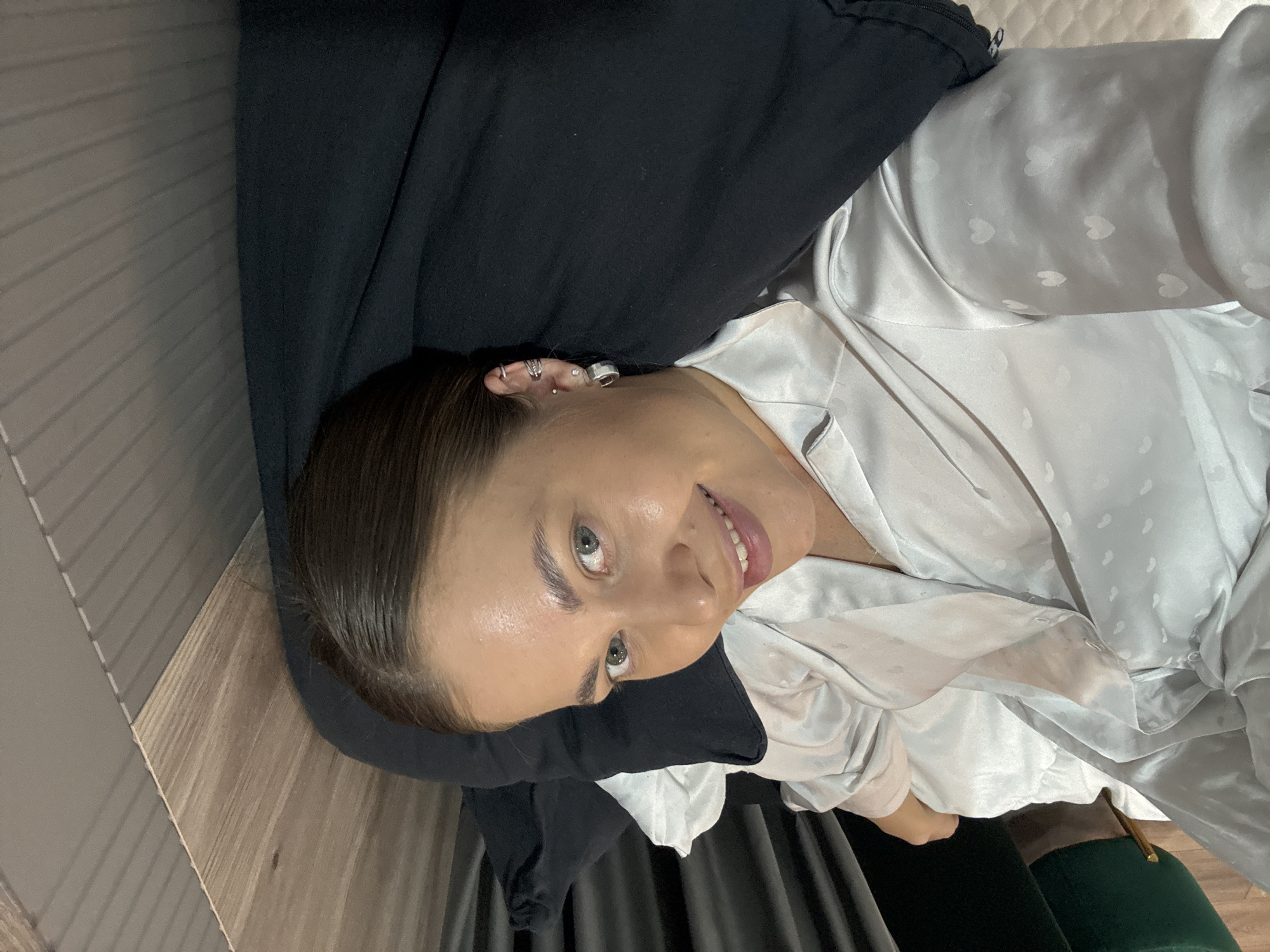 An eye-level shot captures a young woman lying down indoors, looking towards the camera with a gentle smile. Her light brown hair is pulled back, revealing her face with light-colored eyes and well-defined eyebrows. She appears to be in her 20s or 30s, exhibiting a calm and content expression.
She is dressed in a light blue-grey, satin-like shirt or pajama top featuring a subtle, embossed heart pattern. Her left ear is adorned with several silver-toned earrings, including multiple hoops, studs, and an ear cuff. Her head rests on a dark, possibly black, fabric, likely a pillow or blanket.
The background is softly out of focus, depicting an indoor setting. To the left, a patterned wooden surface, possibly flooring or wall paneling, is visible. Behind her, a dark grey, textured fabric, potentially a curtain or upholstery, can be seen. A light-colored, quilted surface, suggesting a duvet or headboard, is also present near her right side. The lighting is soft and even, indicating an indoor environment. The image suggests a relaxed, candid moment within a domestic space, potentially in Chernushka, Russia. No text is visible in the image.