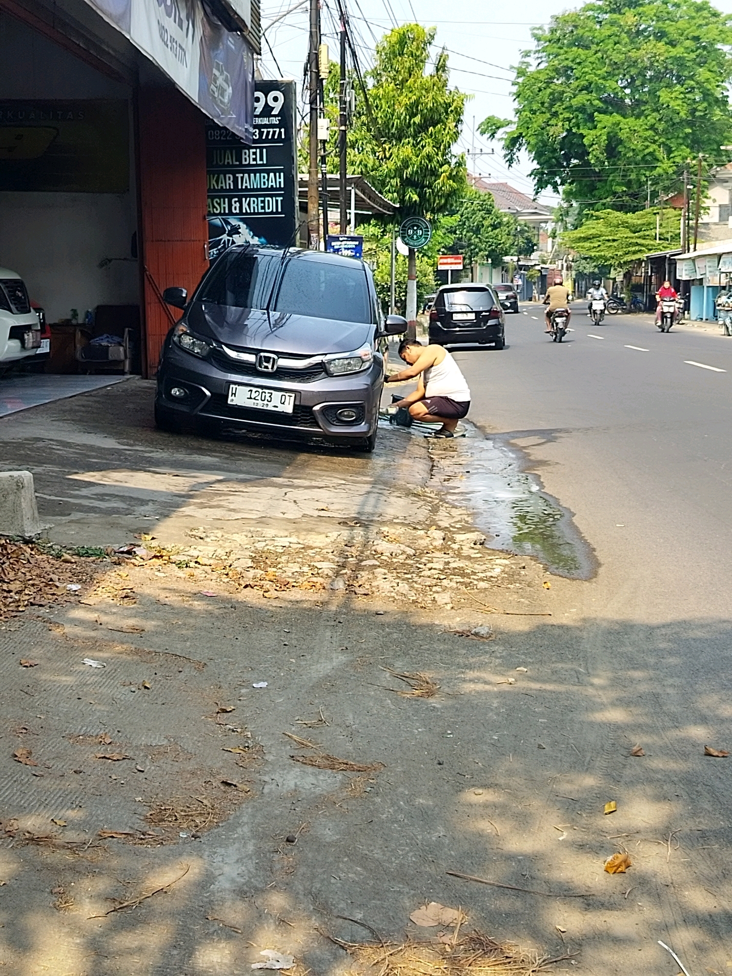 A man wearing a white tank top and dark shorts is squatting on the side of a street in Mojokerto, Indonesia, washing a dark grey Honda Mobilio. The car is parked on a concrete driveway, partially in a garage-like structure. The man is using a green bucket and a white cloth. Water and soap suds are visible on the ground around the car. The street is paved and appears to be a busy road, with several motorcycles and cars in the background. Trees line the street, and buildings are visible in the distance. A sign with "99" and "JUAL BELI TUKAR TAMBAH CASH & KREDIT" is displayed on the building to the left of the car. The scene is illuminated by bright sunlight, casting shadows on the ground.