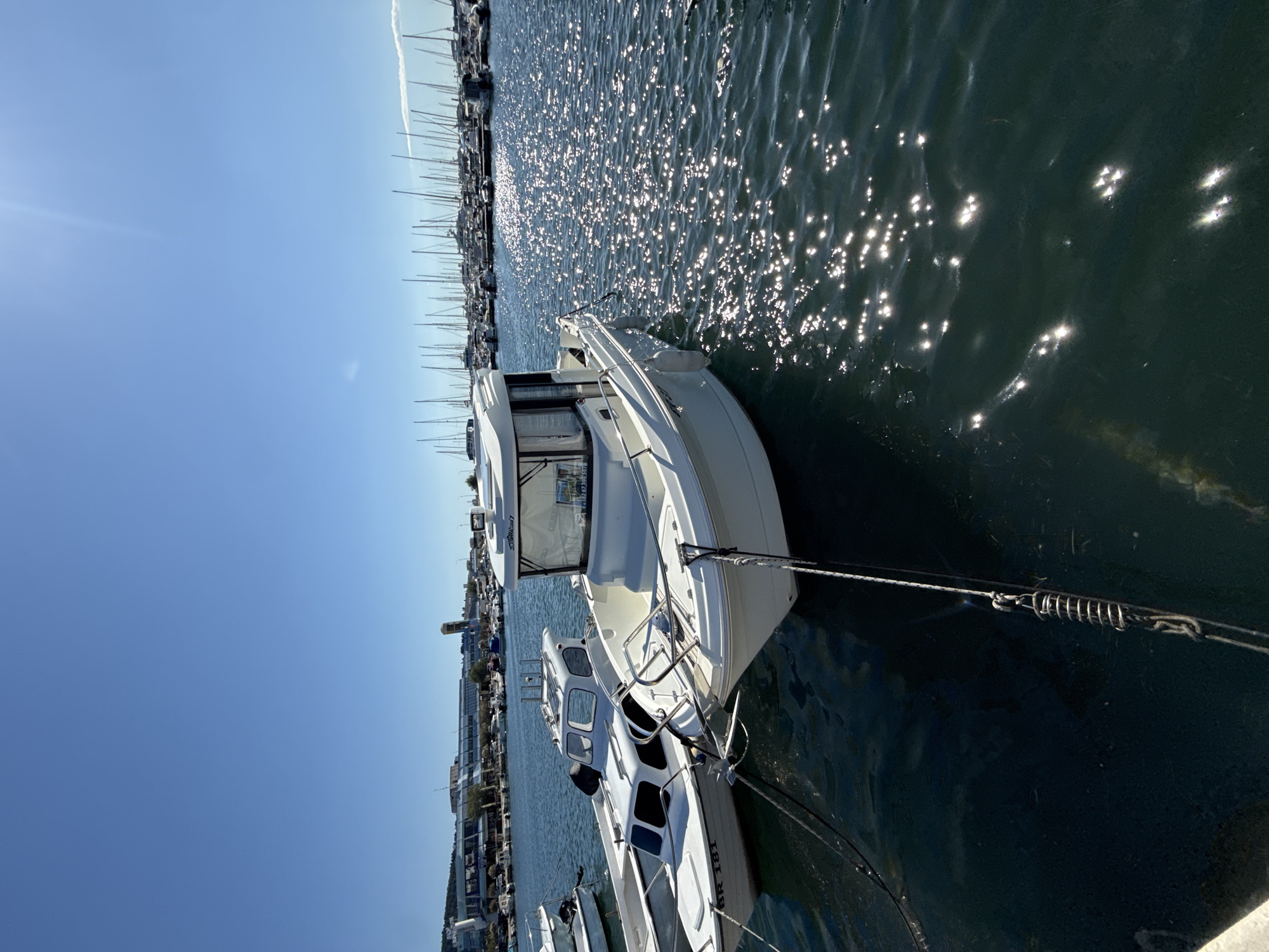 The image shows a bright, sunny day at a marina in Bar, Montenegro. Several white boats are moored in the clear, dark blue water. The foreground is dominated by two white boats, one slightly larger than the other, with cabins. The larger boat in the foreground has a windshield with an inscription that appears to be a phone number. Both boats are secured by ropes to the dock. The water reflects the bright sunlight, creating many sparkling highlights. In the background, a large number of sailboats with tall masts are docked, indicating a busy marina. Further in the distance, a waterfront area with buildings and a tower is visible. The sky is a clear, vibrant blue with a hint of sun flare in the upper right corner. The scene depicts a peaceful and scenic view of a maritime setting.