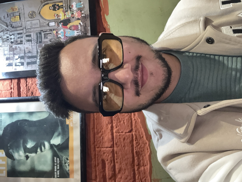 The image features a close-up portrait of a man, taken indoors, likely a selfie. He is positioned slightly off-center to the right, looking directly towards the camera with a neutral or slightly smiling expression. The man has dark, short hair, prominent eyebrows, a mustache, and a neatly trimmed beard. He is wearing large, black, thick-framed glasses with amber or yellow-tinted lenses, which reflect ambient light sources.
He is dressed in a ribbed light blue or teal long-sleeved top, layered under a light beige or off-white jacket, possibly a bomber or varsity style, with some darker stitching or a patch partially visible on the right side of the jacket.
The background suggests an indoor setting, possibly a casual establishment like a cafe or bar. A section of a reddish-brown brick wall is visible behind him, particularly on the lower left and extending upwards behind his head. Above the brick wall, a lighter-colored wall, possibly painted green or a muted tone, is visible.
Two framed items are visible on the wall behind the man. On the upper left, there is a piece of art depicting an urban scene with an archway, buildings, and several cartoonish figures, some appearing to be children. On the lower left, another framed item resembles a vintage magazine cover or poster. This poster features a black and white image of a man's face in profile. Visible text on this item includes: "LIFE", "SETS CHILDREN ADRIFT", "MARLON BRANDO AS ANTONY IN MOVIE, JULIUS CAESAR", "APRIL 20", and a small word "OASIS". The overall lighting appears to be indoor ambient light, creating reflections in the man's glasses.