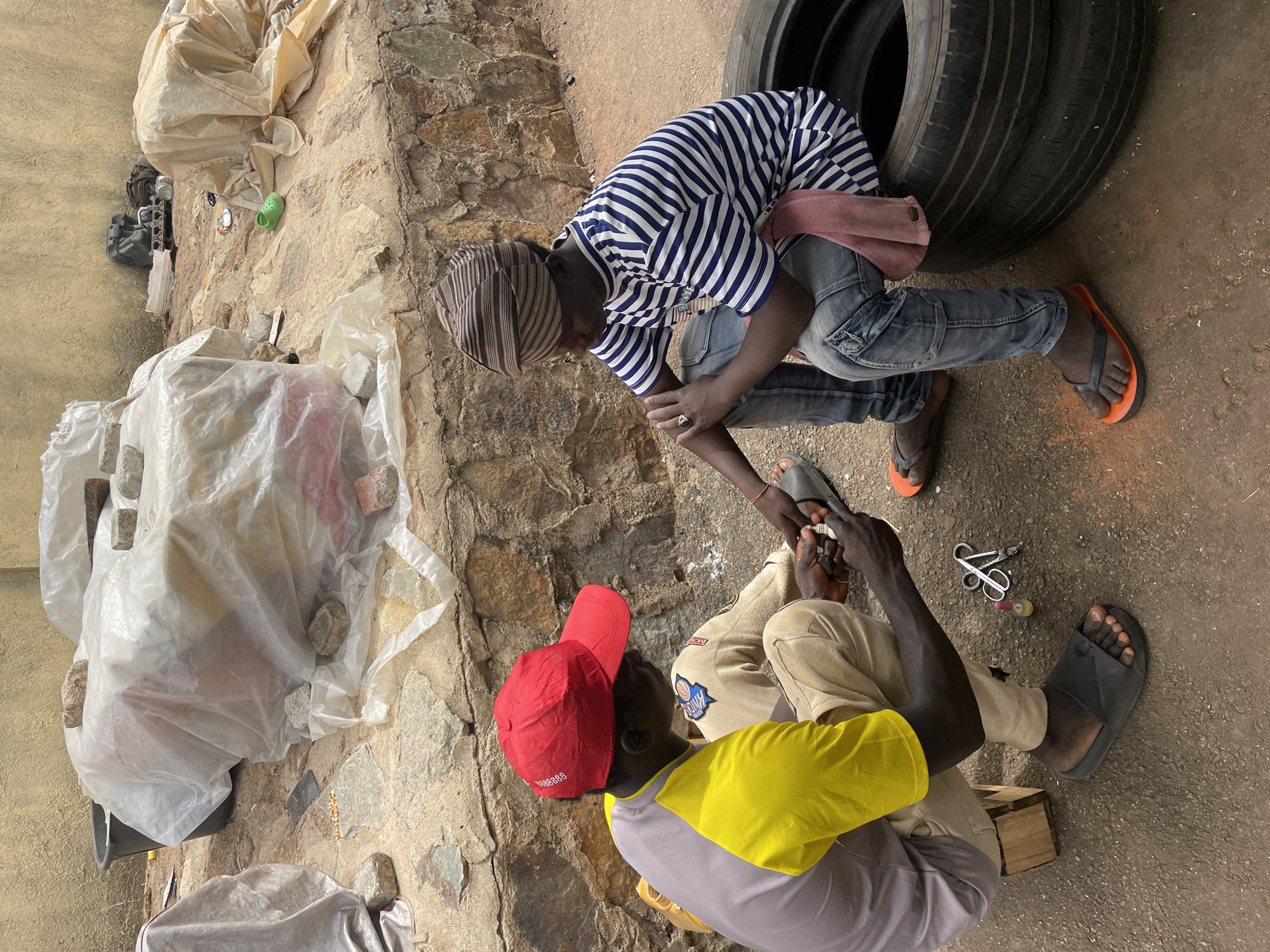 Two men are engaged in an interaction outdoors, possibly repairing something. One man, wearing a striped shirt and a head covering, kneels next to a stack of tires. The other man sits on the ground, wearing a yellow and gray shirt and a red cap, with tools nearby. The location appears to be a roadside in Bukuru, Nigeria, with bags of materials, some held down with rocks, placed next to a stone wall.