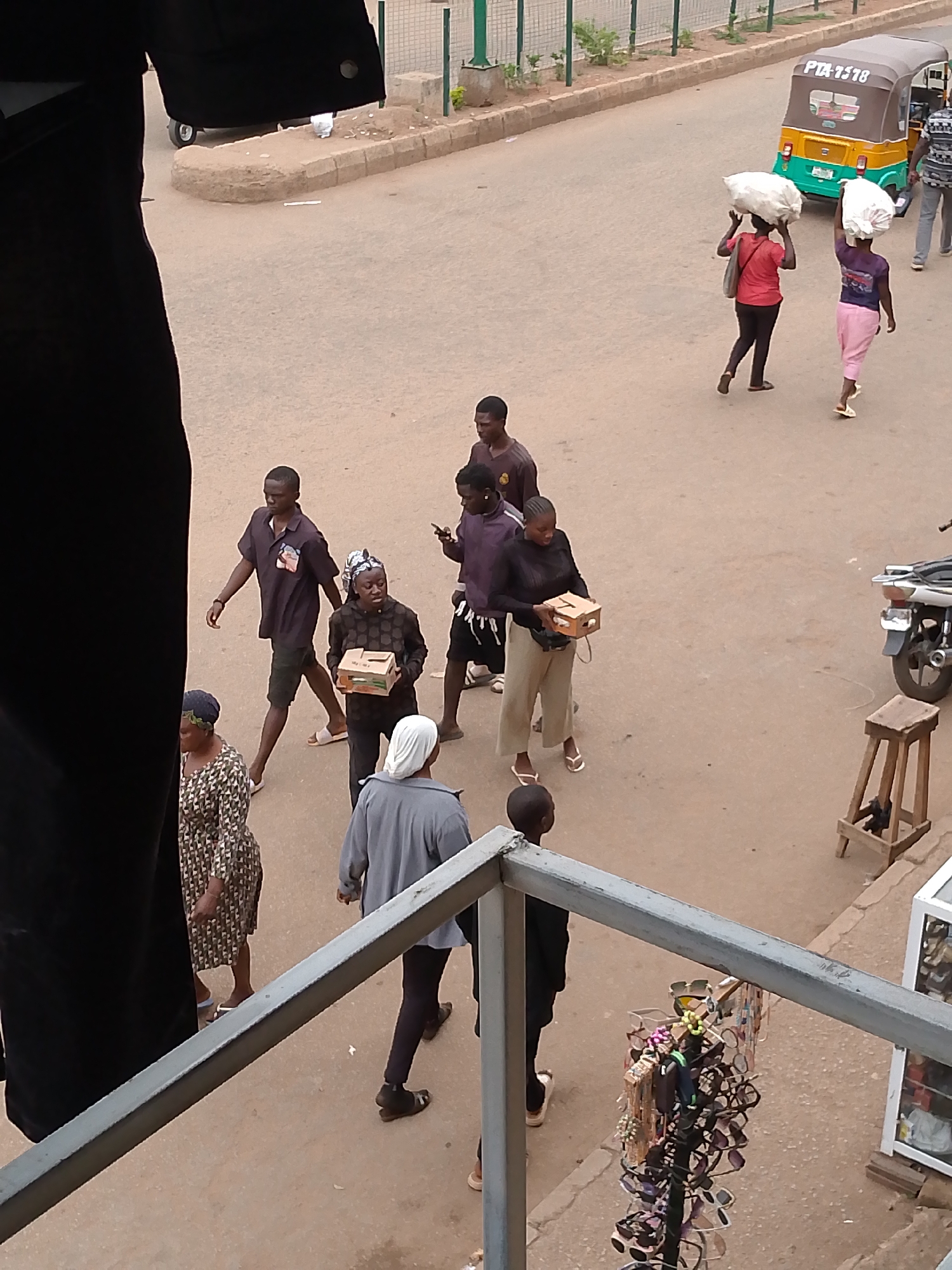 The image shows a street scene in Bukuru, Nigeria. Several people are walking on the street, including two women carrying large white sacks on their heads, and others carrying small boxes. A yellow and green three-wheeled vehicle ("keke") is parked on the road. A motorcycle is parked on the side of the road, near a wooden stool. The view is from an elevated perspective, looking down onto the street. A dark-colored garment is partially visible on the left side of the frame. A metal railing and a display of sunglasses are visible in the foreground.