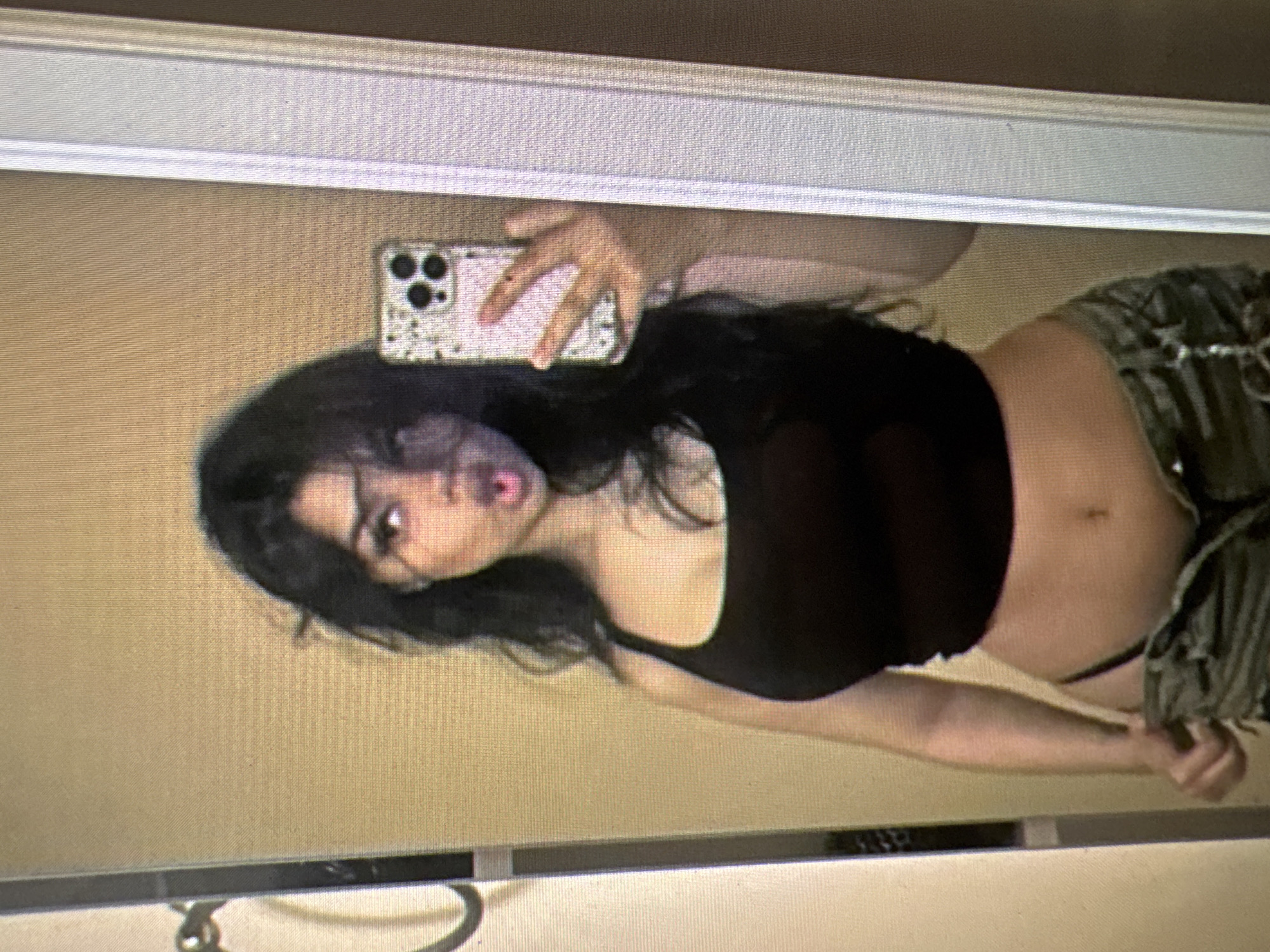The image shows a young woman in a black tank top and camouflage pants, holding a phone to take a mirror selfie. She is standing in front of a plain wall, with a mirror on her right and a door frame on her left. The setting appears to be indoors, likely a bedroom or bathroom, but there are no specific elements in the image that would allow for identification of the location within the city of Castelo Branco, Portugal.