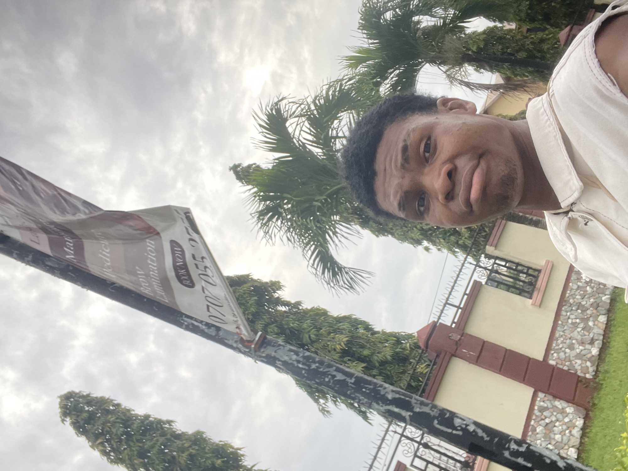 A young Black male is taking a selfie outdoors in Moba, Nigeria, with a slight frown. He is wearing a light-colored, sleeveless collared shirt. In the background, a tall, dark pole with a banner attached is visible, displaying text such as "Manicure," "Pedicure," "Brow Lamination," and a phone number, "0707 055 2". The banner appears to be advertising beauty services. Behind the pole and banner, a wall with stone and plaster accents, a black metal fence, and lush green trees, including palm trees, are present. The sky is overcast with grey clouds. The overall scene suggests an outdoor setting in a residential or commercial area in Nigeria.