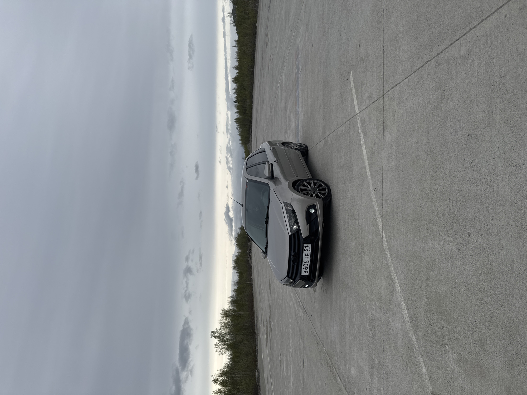A gray Lada car is parked on a gray concrete surface, possibly a parking lot or an open area. There is a line marked in white on the concrete, indicating a parking space. A forest can be seen in the background, and the sky is cloudy and overcast. The car's license plate reads "B606HE 51," which indicates it is registered in the Murmansk region of Russia. The image was likely taken near Kirovsk, Russia.