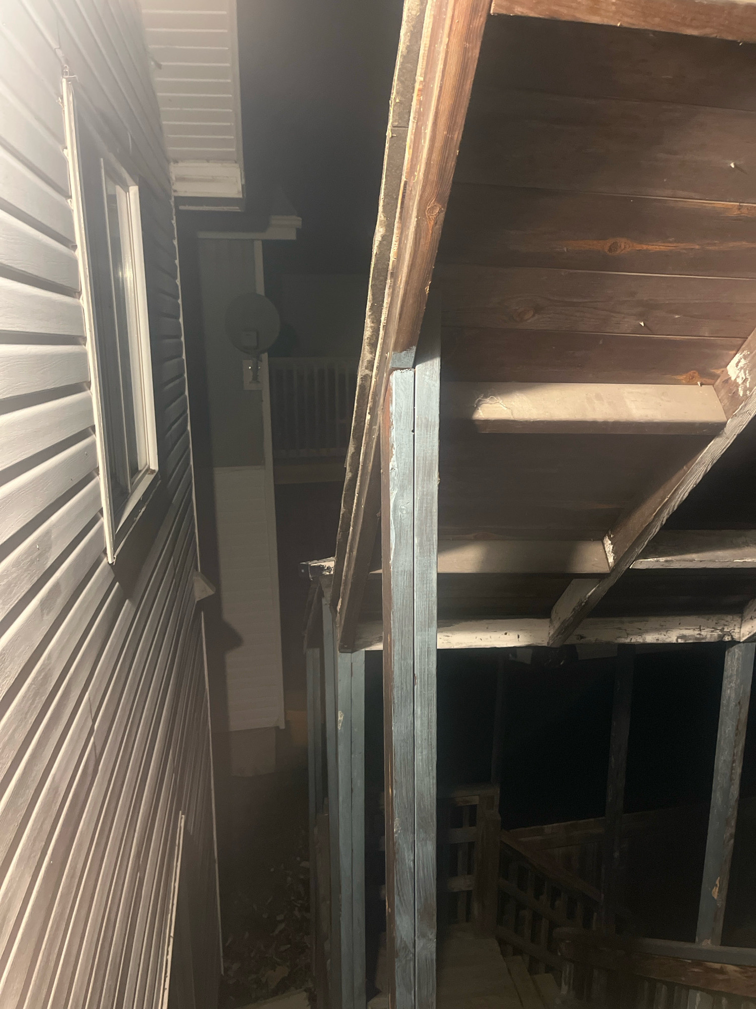 The image depicts an exterior view of a building at night, possibly a residential property. On the left, a section of siding, likely vinyl, is visible. The siding is light-colored and appears to be in good condition, with a window set into it. Adjacent to the siding is a dark, covered porch or walkway, supported by wooden posts. The underside of the porch roof is made of wooden planks. The porch leads to a set of stairs, which descend into darkness. A satellite dish is mounted on the side of the building in the background. The scene is dimly lit, with the primary light source appearing to be artificial and directed towards the building. There are no people or specific actions depicted in the image. The location is identified as Temiskaming Shores, Canada.