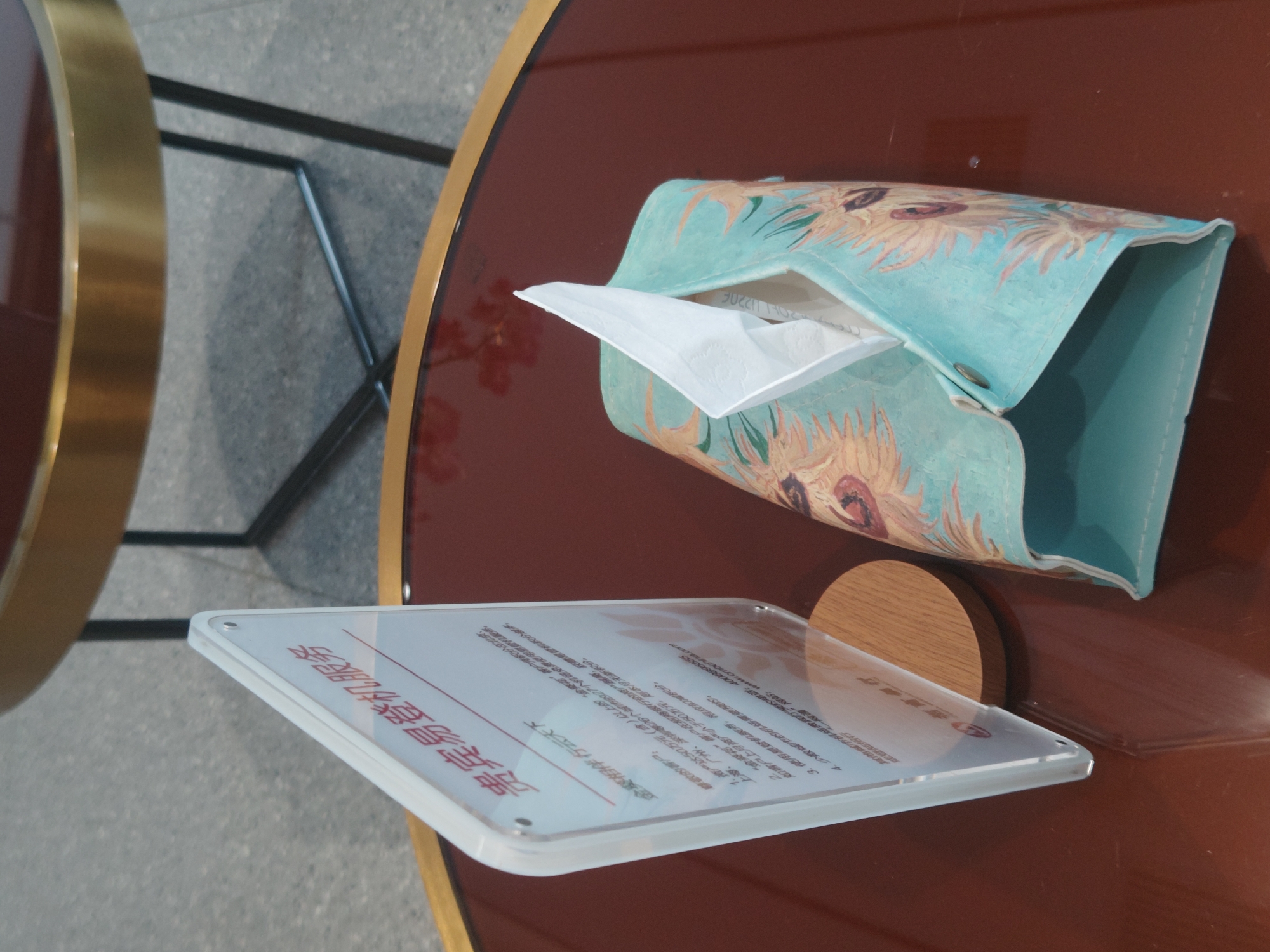 An overhead shot shows a table with a clear acrylic sign and a tissue box. The sign displays Chinese text and is supported by a wooden base. The tissue box, adorned with a sunflower pattern, sits to the right of the sign and contains a white tissue peeking out. The table's surface is a dark, glossy red, reflecting the objects. In the background, a portion of another round table with a gold rim and black metal legs is visible, suggesting an indoor setting. The image is likely taken in a public space within the Chongqing High-tech Zone, China, possibly a lounge area or reception.