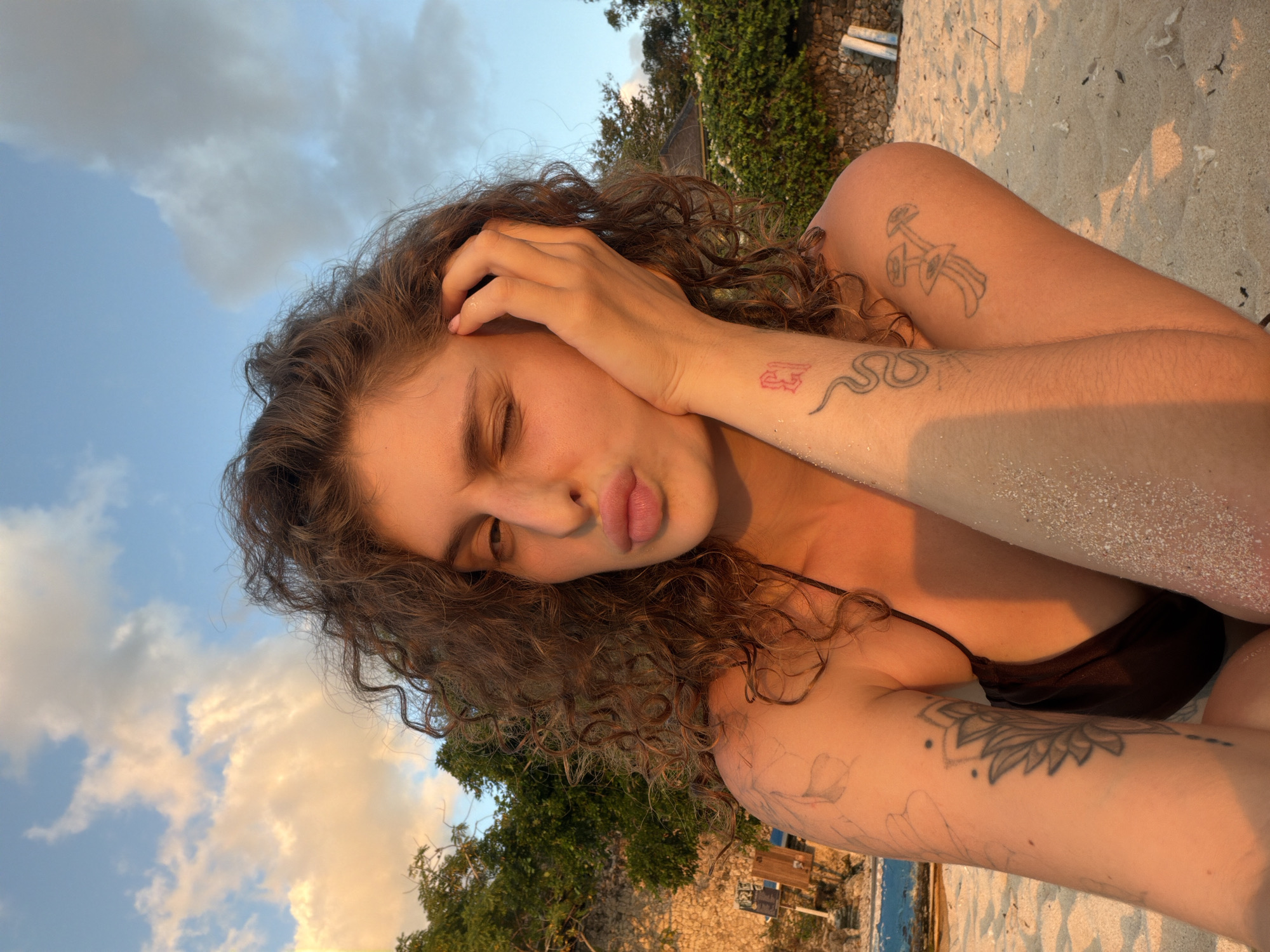 A medium shot captures a person with curly brown hair on a sandy beach during golden hour. The person is wearing a dark bikini top and is partially submerged in sand up to their knees. Their right arm is raised, resting their head against their hand, while their left arm is extended, showcasing tattoos of a lotus flower, a snake, and mushrooms. They are winking and puckering their lips, with the sun casting a warm glow on their skin. The background features lush green trees and a stone wall, suggesting a tropical or coastal environment. The sky is a clear blue with scattered clouds tinged orange by the setting sun. The sand is light beige and speckled with fine white grains.