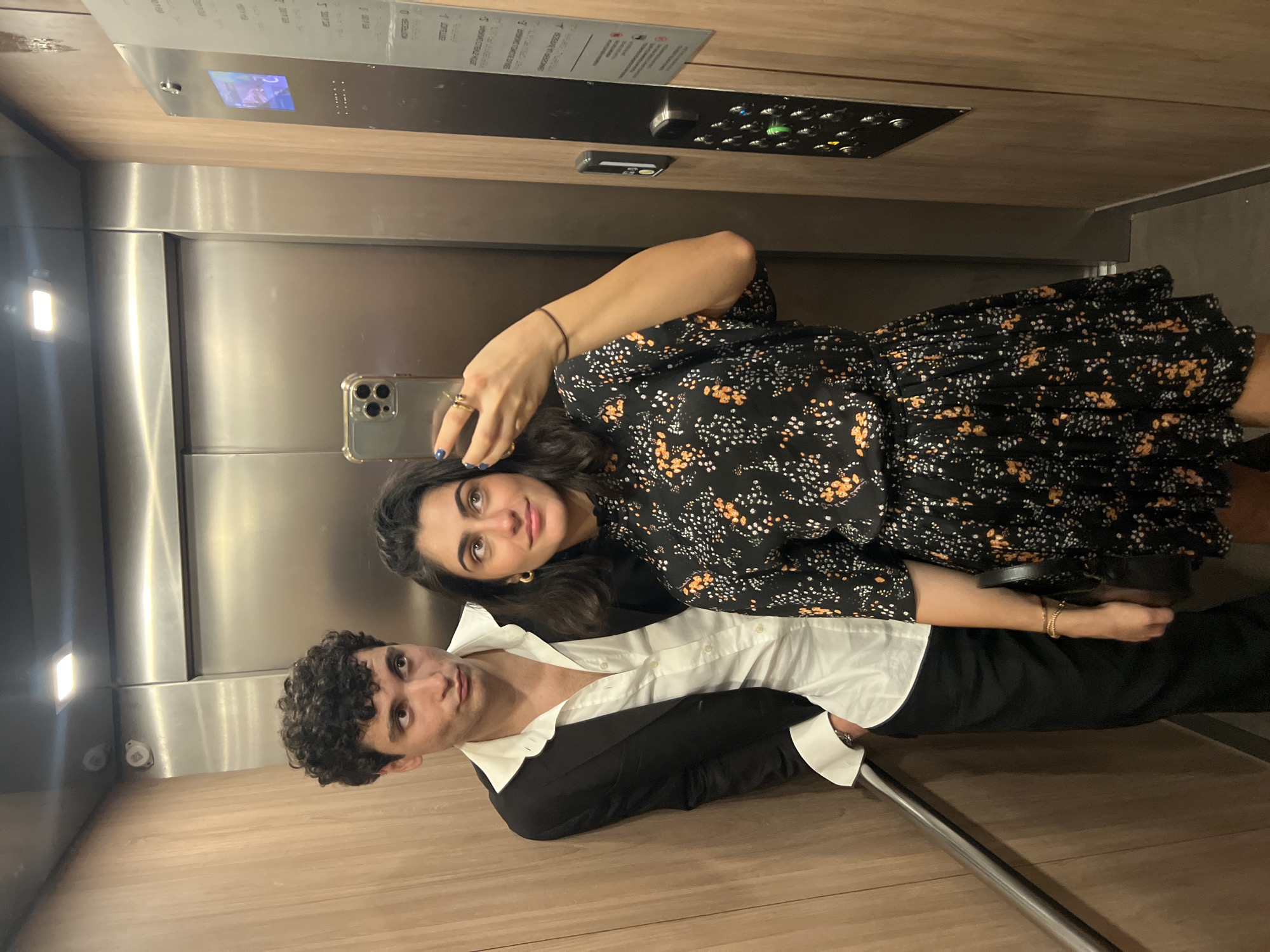 A young couple is taking a mirror selfie in an elevator with wood-paneled walls. The man on the left has curly dark hair and is wearing a white dress shirt with an unbuttoned black blazer and black pants. He is leaning against the wall. The woman on the right has dark hair and is wearing a black floral print dress. She is holding her phone up to take the photo. Her left hand, adorned with rings, is adjusting her hair. The elevator doors are to the right, and an elevator control panel with buttons is visible on the wall. The background is an elevator interior. The location is an unspecified elevator in Paris, France.
