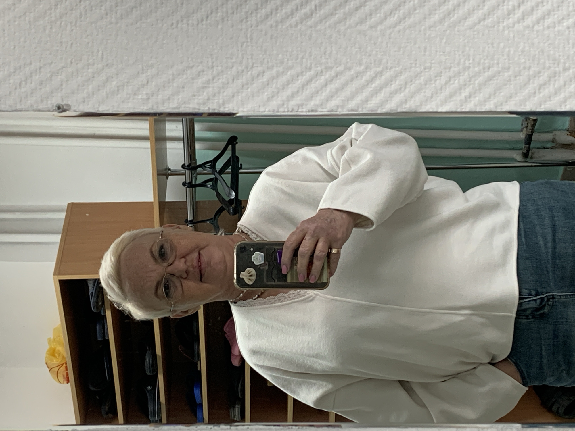 A selfie taken in a mirror features an older woman with short white hair, wearing glasses, a white sweater with lace trim, and jeans. She holds a smartphone in her hand. Behind her is a light brown wooden shelf with various dark-colored objects inside, likely shoes. To the right of the shelf, there is a clothes rack with black hangers and a light green wall with horizontal pipes. Based on previous information provided to the model, the location is in the city of Chaikovsky, Russia.