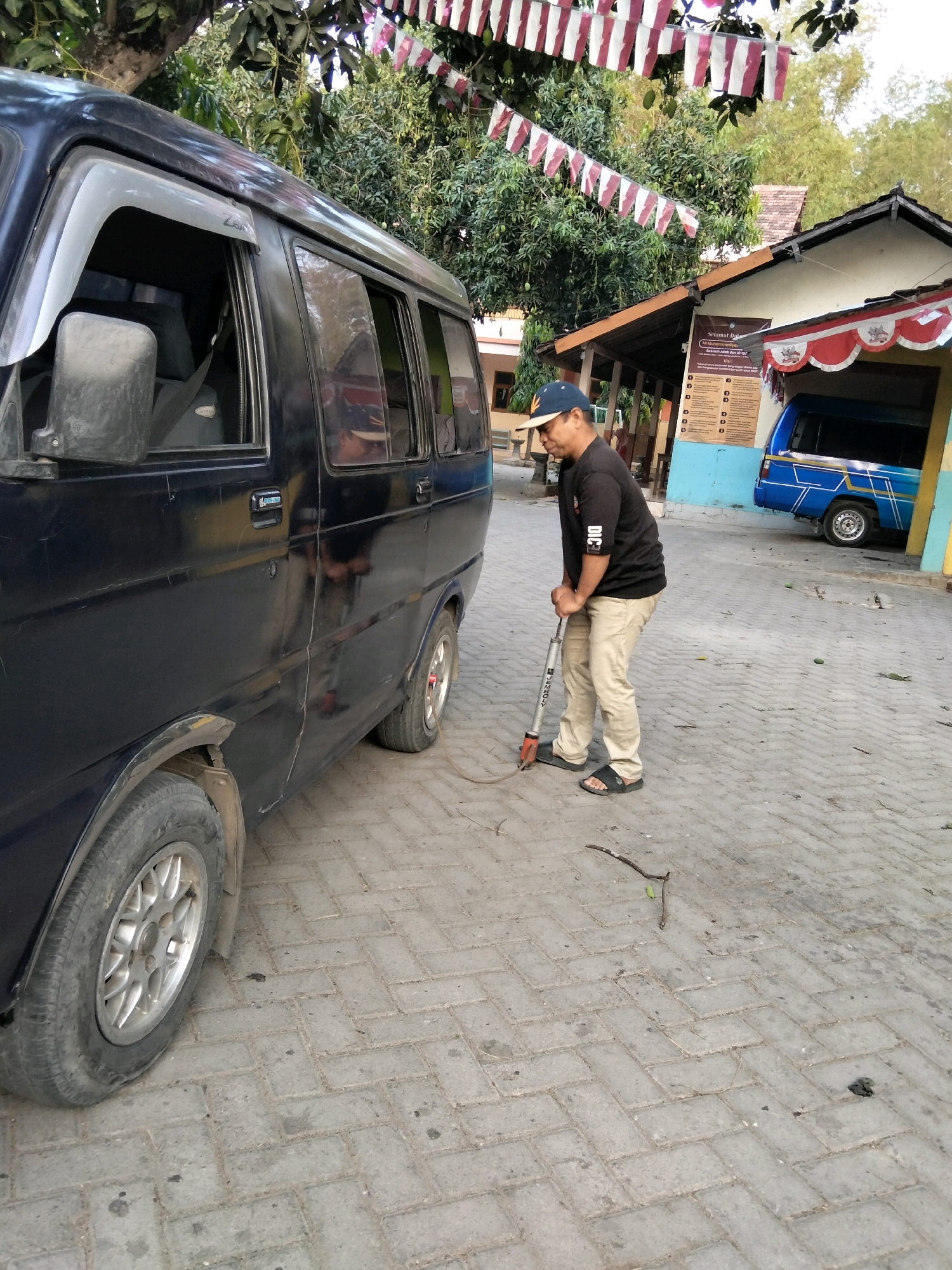 A person in Jenangan, Indonesia, wearing a black t-shirt, tan pants, a blue baseball cap, and black sandals, is standing next to a dark blue van. The person is holding a tire inflator and appears to be in the process of inflating a tire. The van is parked on a brick-paved surface. In the background, there is a building with red and white banners hanging, and trees with fruit are visible. A second blue van is parked in front of the building. The scene appears to be outdoors, possibly in a residential area or a parking lot.