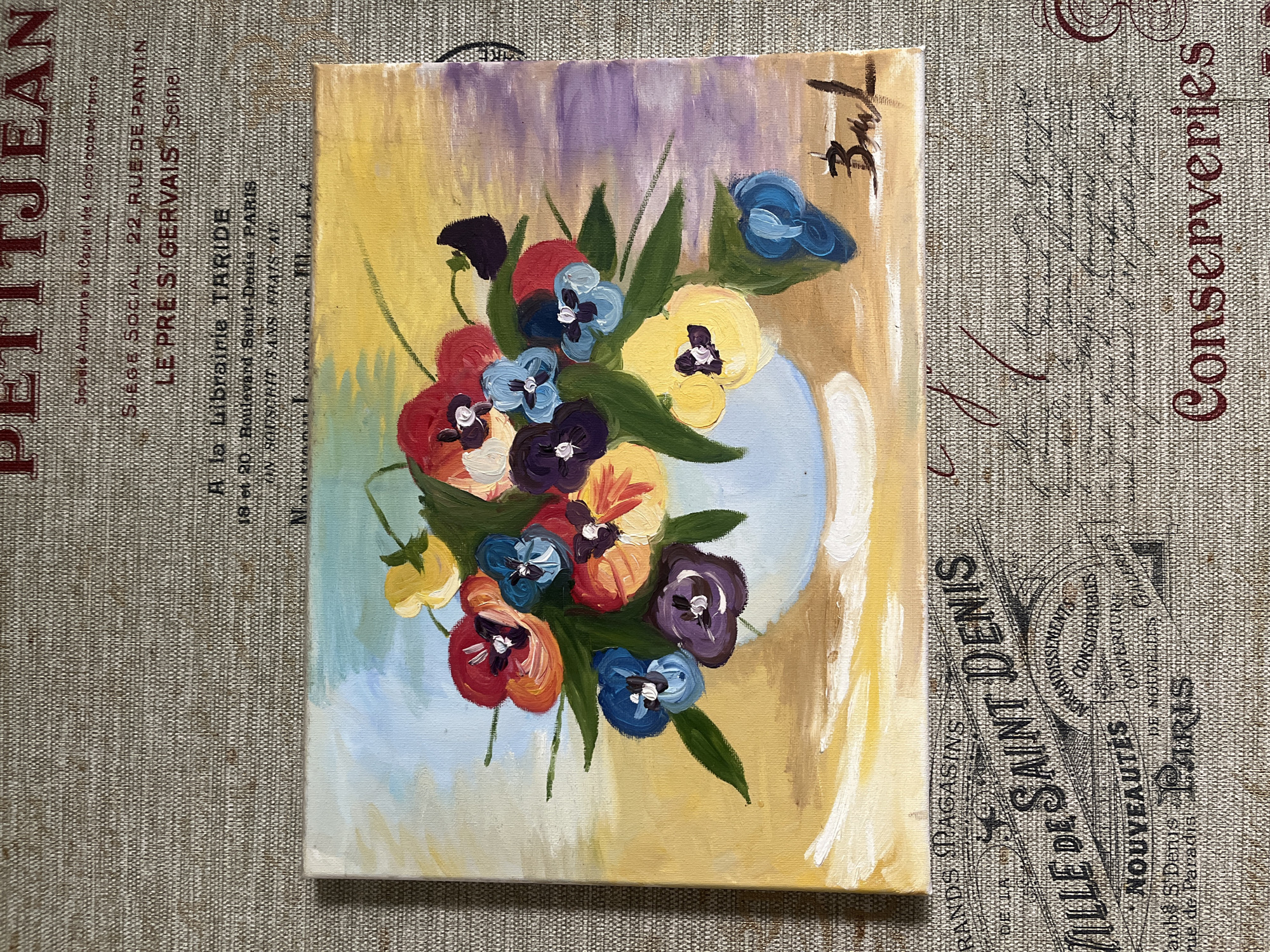 The image shows a small, rectangular painting of a bouquet of pansies in a light blue vase. The flowers are a mix of colors: red, blue, yellow, orange, and purple. The background of the painting is a blend of yellow, brown, and purple hues. The painting is signed "BML" in the upper right corner. The painting sits on a patterned fabric or tablecloth. The fabric is printed with French text, including phrases like "PETITJEAN," "LE PRE ST GERVAIS (Seine)," "Boulevard Saint-Denis PARIS," "VILLE DE SAINT DENIS," and "Conserveries."