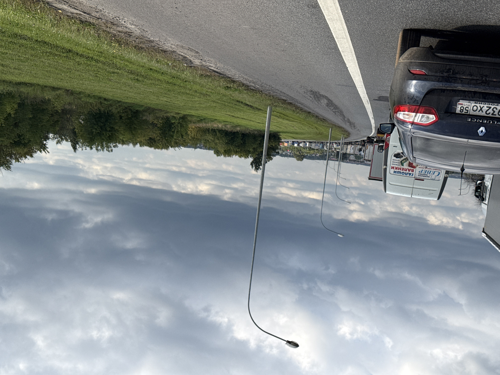 In a city of Unknown, Unknown, this image captures a scene of a traffic jam on a cloudy day. The perspective is from behind a dark-colored Renault Fluence sedan, facing forward into the queue of vehicles. The sedan's rear is prominently featured, with its license plate reading "932 XO 58". To the left of the sedan, a white truck is visible, bearing text that includes "СЕВЕР" and "ГАЛОШИ ВАЛЕНКИ". Ahead of these vehicles, a long line of trucks stretches into the distance, indicating a significant traffic standstill. The road is paved, with a white line marking the lane. On the right side of the road, a grassy embankment slopes upwards, dotted with trees and vegetation. Several streetlights are positioned along the side of the road. The sky is overcast with heavy, dark clouds, suggesting the possibility of rain. The overall impression is one of slow-moving or stationary traffic in a semi-rural or suburban setting.