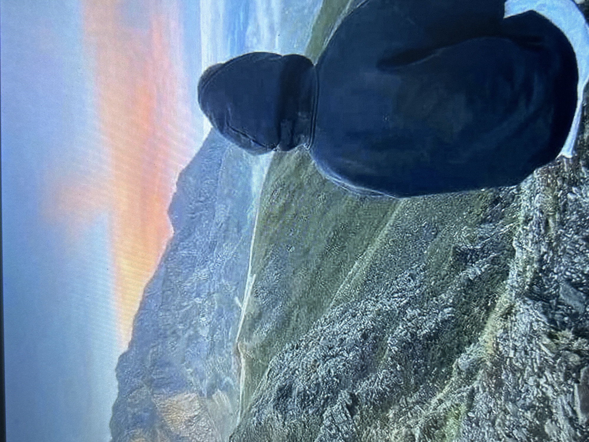 The image captures a solitary figure, viewed from behind, positioned on a rocky, elevated terrain, observing a vast mountainous landscape under a colorful sky, purportedly located in the city of Burgas, Bulgaria. The individual is wearing a dark, possibly black or navy blue, hooded garment, with the hood fully drawn over their head. A strip of white fabric, likely from an undergarment, is visible beneath the lower edge of the dark top. The immediate foreground consists of rugged, greyish-green rocky ground with sparse patches of light vegetation. Extending into the midground are undulating hills covered in a mix of green foliage and exposed rock, with a light-colored, narrow path or track discernible horizontally across the landscape. Further in the background, more imposing, craggy mountains with pronounced ridges exhibit a bluish hue due to atmospheric perspective. The sky transitions from a clear, light blue overhead to vibrant shades of orange, pink, and red closer to the horizon, characteristic of either dawn or dusk. To the far right, a hazy, light blue expanse suggests distant clouds, mist, or a body of water. No other individuals, structures, or objects of note are present in the visible scene. The image exhibits visible screen artifacts, including vertical lines and pixelation, indicating it is a photograph of a digital display.