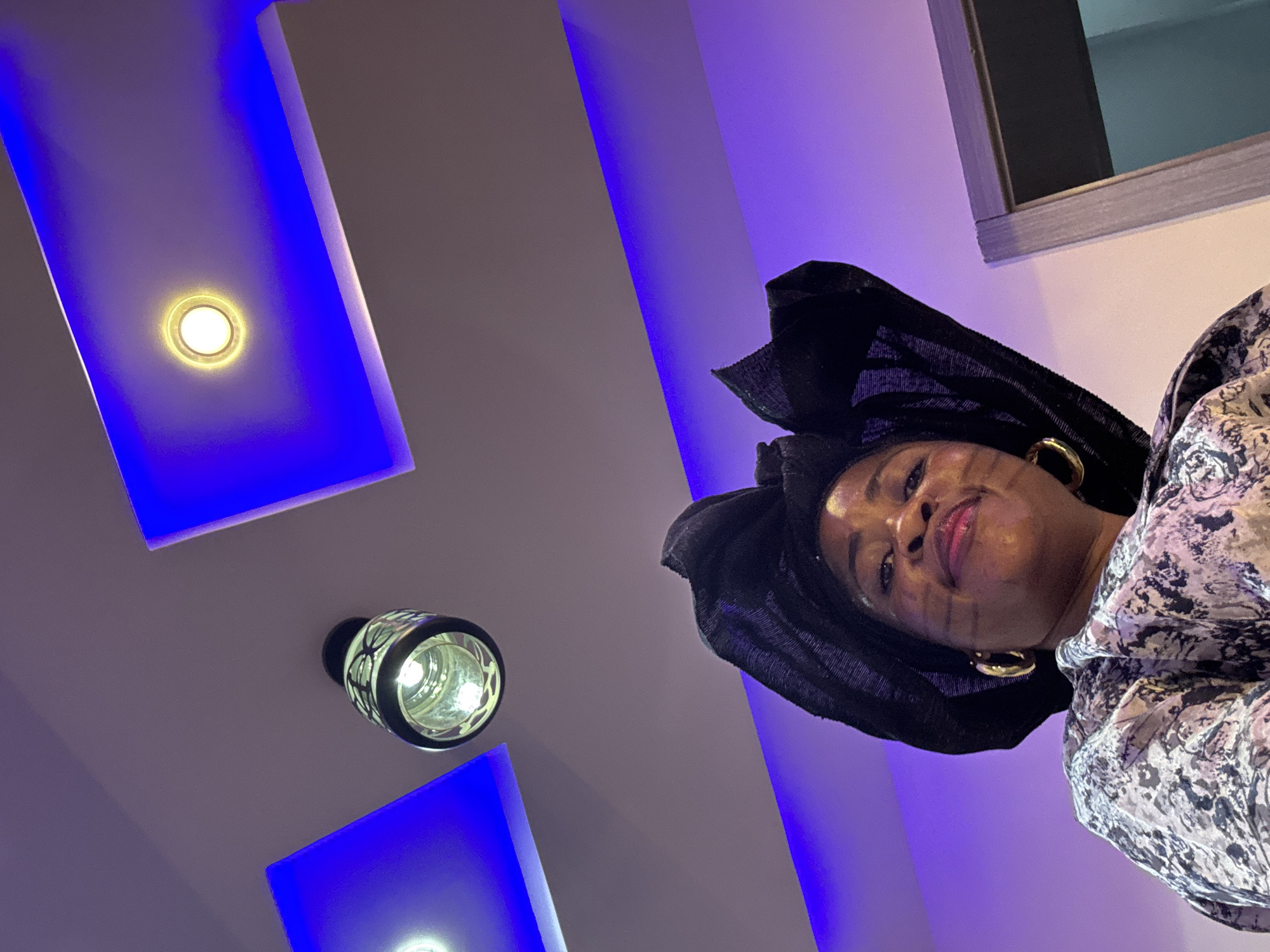 A low-angle shot captures a woman from the chest up, looking towards the camera with a slight smile. She is wearing a black headwrap with intricate folds and large gold hoop earrings. Her attire is a patterned fabric with gray, black, and white designs. The background features a modern ceiling with recessed blue LED lighting and a decorative pendant light fixture. The woman's face has subtle dark markings on her cheeks, possibly traditional face paint or makeup. The scene appears to be indoors, possibly in a residential or event space in Ajara, Nigeria.