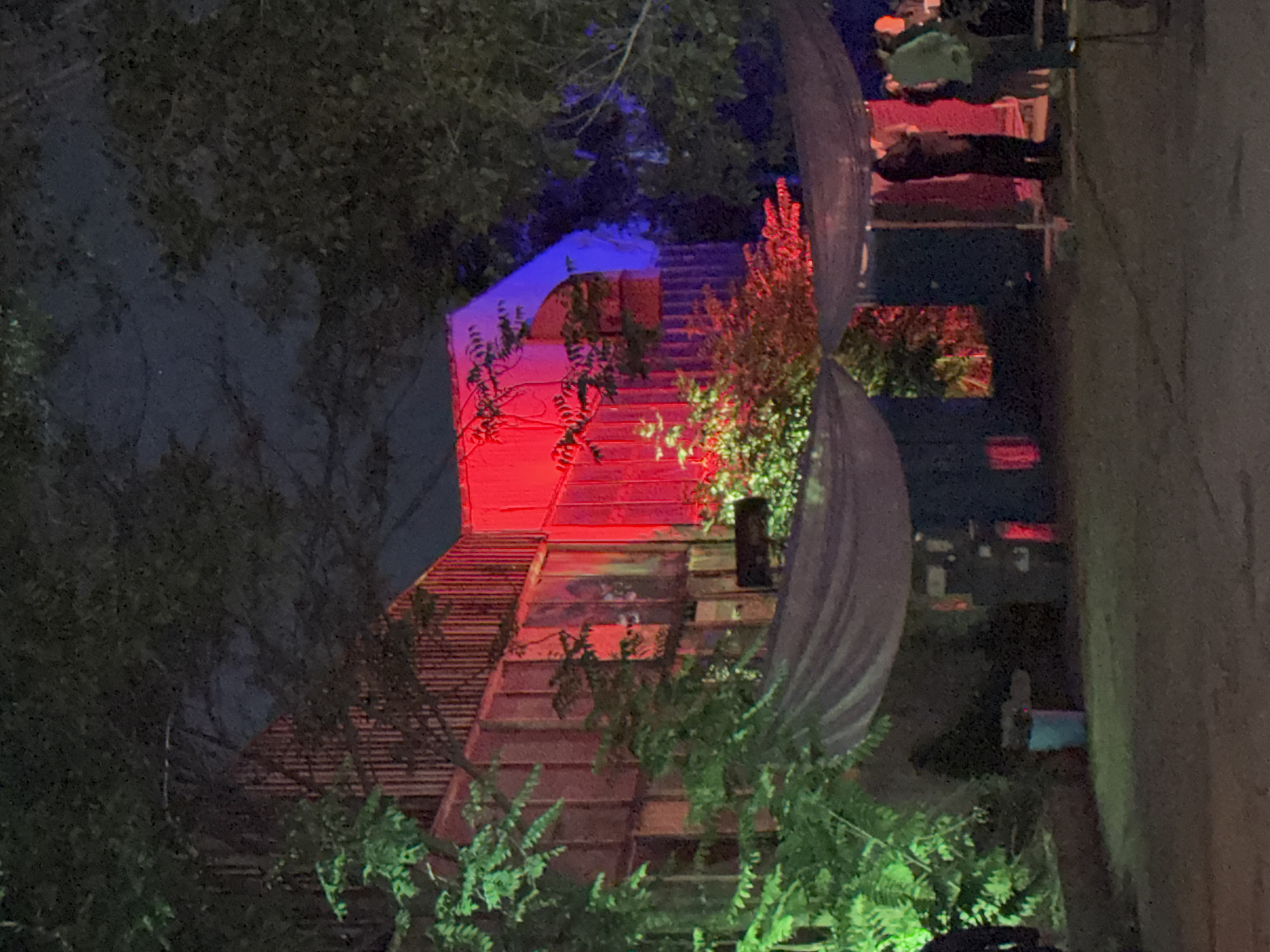 The image depicts an outdoor nighttime scene in Sofia, Bulgaria, likely at a venue or event. A multi-story structure, possibly a building or a temporary stage setup, is illuminated with vibrant red and blue lights. The facade of the structure appears to be made of corrugated metal or wood panels, with vertical lines visible. Trees with lush green foliage are in the foreground and frame the scene.
Beneath the structure, a tarp or awning is stretched out, supported by poles, creating a covered area. Several individuals are visible within this covered space and around the perimeter. Some are standing, seemingly observing or interacting. One person is wearing a light-colored jacket, another is in darker clothing, and two individuals are partially visible in the background.
In the lower left portion of the image, green lighting illuminates more foliage. A light blue trash bin is positioned near the base of the structure on the left. Speakers or sound equipment are also present in the covered area. The ground appears to be unpaved or dirt. The overall atmosphere is characteristic of an outdoor gathering or performance space at night, with emphasis on the dramatic lighting.