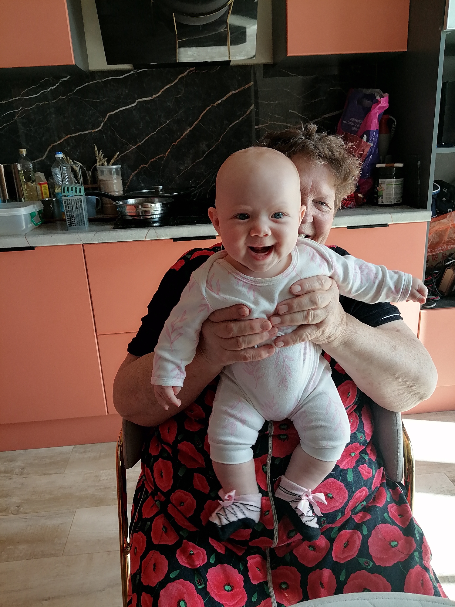 In a brightly lit kitchen, an older woman is holding a smiling baby. The woman has short, curly brown hair and is wearing a black shirt with red floral patterns. The baby is bald and wearing a white onesie with a pink leaf pattern and pink socks with bows. The baby is looking directly at the camera with a wide, joyful smile, and the woman is also smiling, looking slightly off-camera. They are seated in a chair with a metallic frame, and the background shows kitchen cabinets in a peachy color and black marble-patterned backsplash. On the counter, there are bottles, containers, and cookware, suggesting a domestic setting. A colorful bag is visible on a shelf behind them, and the overall impression is one of warmth and familial affection. The image appears to be taken during daylight hours, with ample natural light illuminating the scene. The floor is made of light-colored wood planks. No visible text is present in the image.