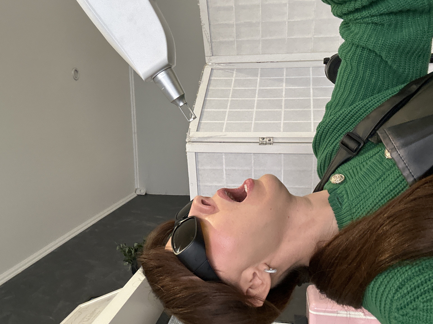 The image, taken in Riga, Latvia, depicts a woman undergoing a cosmetic or medical procedure in an indoor setting. The woman is shown from her upper torso, lying supine and looking upwards with her mouth agape. She has brown hair, fair skin, and is wearing dark protective goggles that cover her eyes. She sports a green knitted top or cardigan with distinct golden buttons and a black strap crossing her shoulder. A small, sparkly hoop earring is visible in her right ear.
A white, handheld treatment device, likely a laser, with a silver tip, is positioned directly above her face, suggesting a procedure is actively taking place or about to begin. In the background, light gray walls and a dark gray floor are visible, bordered by a white baseboard. A white, decorative trunk or chest with a grid pattern is situated behind her head, and a small, partially visible pink object (possibly furniture) is in the lower right corner. A small plant in a pot can be faintly seen on the floor. The overall ambiance suggests a professional clinic or salon environment. No clear text is discernible on any objects. The woman's expression, with her mouth open and eyes protected, conveys a sense of passive participation in the treatment.