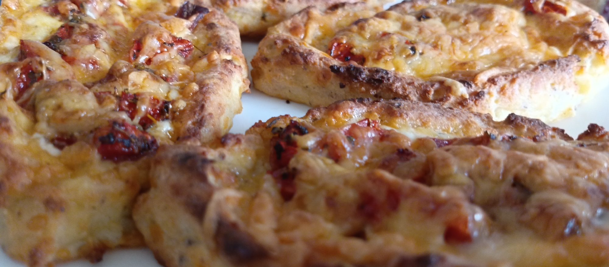 The image presents a close-up view of several small, round, savory pastries or mini pizzas. Each item features a golden-brown, baked crust, suggesting a flaky or slightly crispy texture. They are generously topped with melted, bubbly cheese, which exhibits varying shades of yellow and orange, with some areas appearing slightly browned and glossy.
Scattered across the cheese are pieces of red topping, most likely tomatoes, some of which show signs of being caramelized or roasted. Dark specks, possibly dried herbs like oregano or basil, are also visible within the melted cheese and among the tomato pieces, adding to the visual texture.
The pastries are arranged closely together on a plain, white surface, which could be a plate or serving platter. Both the foreground and background of the image are softly blurred, creating a shallow depth of field that keeps the focus sharply on the food. The lighting is bright and even, highlighting the textures and colors of the baked goods, making them appear appetizing. There are no people, visible text, or specific location cues beyond an indoor setting. No particular activity is taking place; the food is simply presented.
