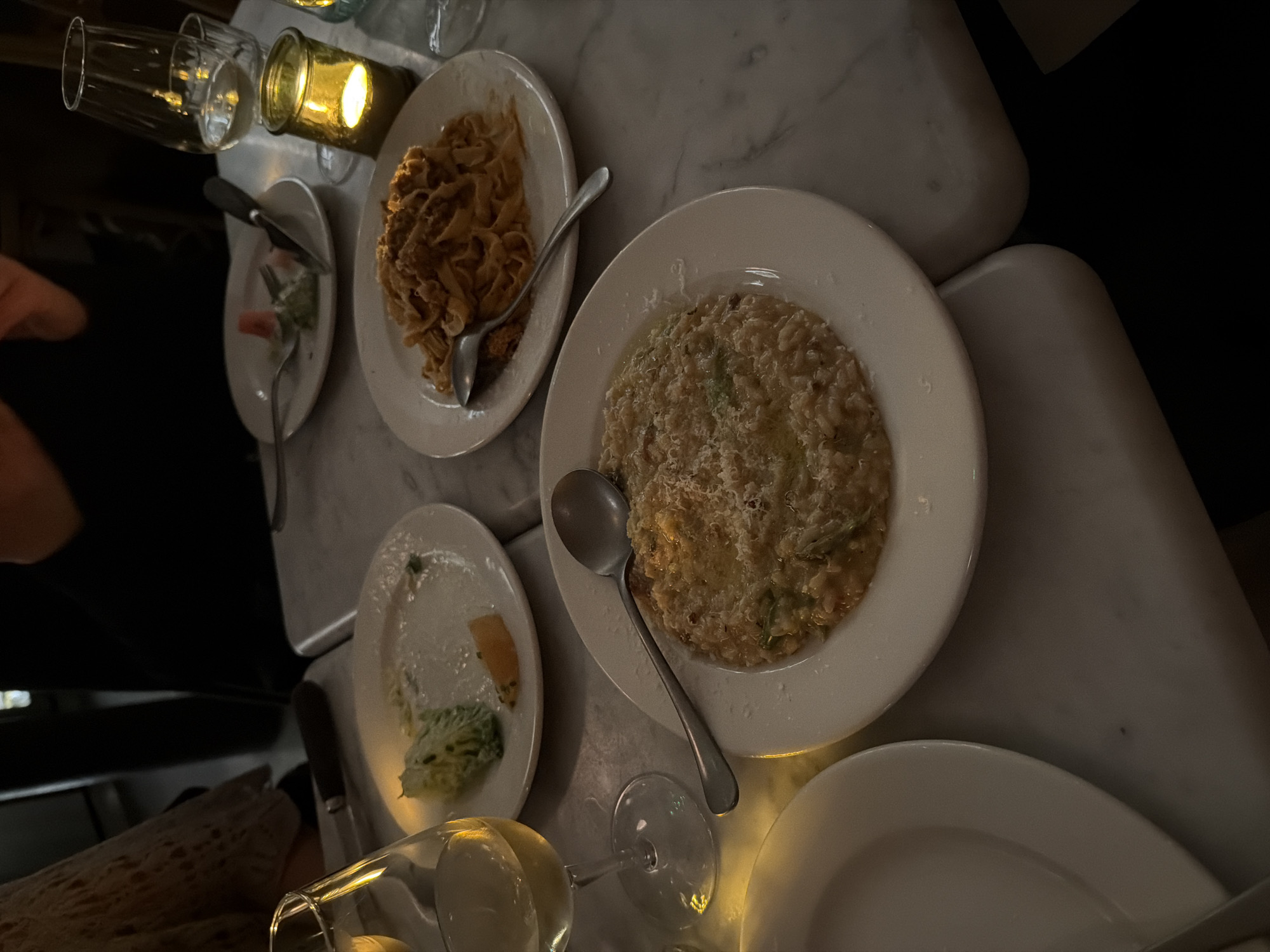 The scene is a dimly lit restaurant interior, possibly in London, United Kingdom, based on the style and potential clientele. The focus is on a marble-topped table cluttered with several plates of food, some nearly finished, and drinkware.
Objects:
* Plates: Four visible plates. One with remaining fettuccine pasta, one with risotto, and two with leftover salad and vegetables.
* Cutlery: Forks and spoons are present, used and placed on the plates.
* Glassware: Several wine glasses, some empty, and a candle holder.
Food:
* Fettuccine pasta.
* Risotto.
* Salad and vegetables.
Environment:
* Dimly lit, warm lighting suggesting a relaxed atmosphere.
* Marble tabletop.
* Indicates an intimate setting.
People:
* Partial view of people possibly seated at the table.
Actions/Interactions:
* Evidence of a meal consumed.
* Conversation.
Overall impression: It suggests a casual dining experience.