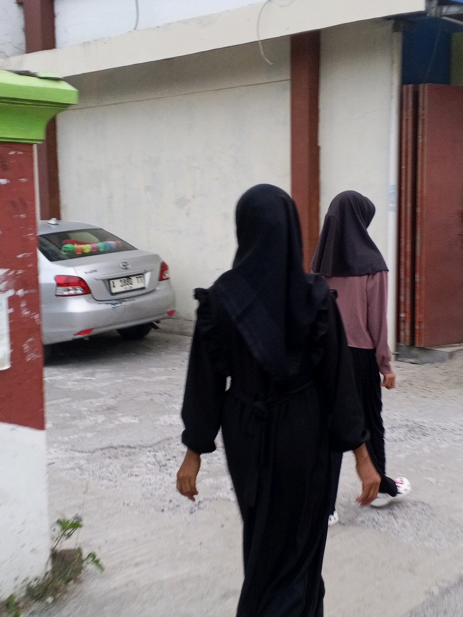 Two women, one in a black hijab and abaya and the other in a dark headscarf and a light purple long-sleeved top and black skirt, walk away from the camera on a paved outdoor area. The woman in front is wearing a black abaya with ruffled sleeves and a sash tied at the waist. The woman behind her is wearing a dark headscarf over a light purple top and a black skirt. She has white and pink patterned shoes. To the left of the women, a silver sedan is parked. The car's license plate reads "A 2880 TTT." Colorful items are visible on the rear windshield shelf of the car. The background consists of a building with a cream-colored wall, red vertical beams, and a white overhang. A red and white striped concrete pillar is visible on the far left of the image. Small green plants are growing from cracks in the ground near the pillar. The overall environment appears to be an outdoor public space, possibly a street or parking lot, in a city like Ciruas, Indonesia.