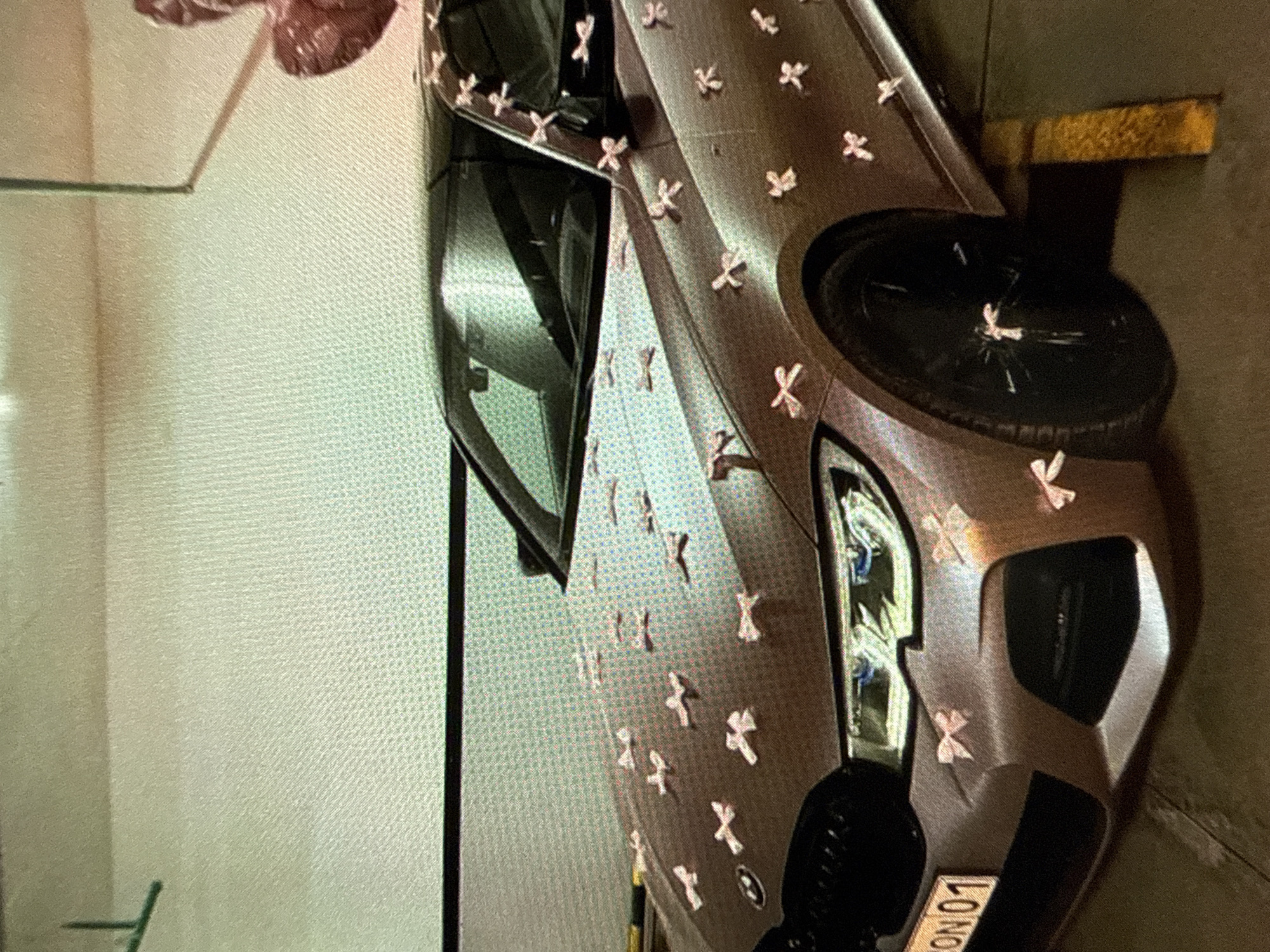 A silver BMW is adorned with small, light-pink ribbon bows. The car has balloon decorations attached. The location is a parking garage, location unknown.