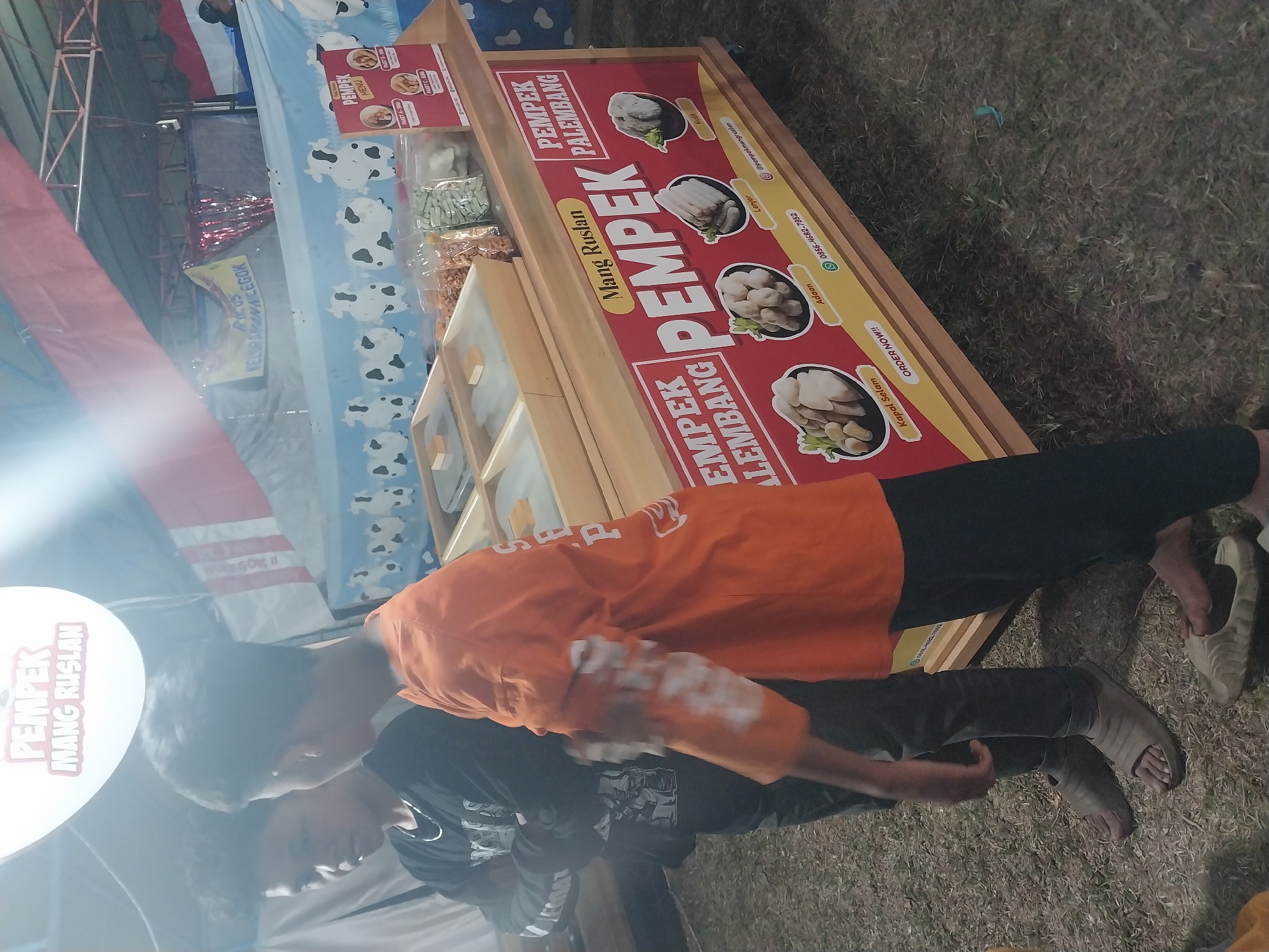 The image depicts an outdoor food stall with a red and yellow banner advertising "PEMPEK PALEMBANG" from "Mang Ruslan." The stall displays various types of pempek, a traditional Indonesian fish cake, with illustrations of "Kapal Selam," "Adaan," "Lenjer," and "Kulit." A phone number and Instagram handle are visible on the banner. Two young boys are standing near the stall, with one in the foreground wearing an orange shirt and the other in the background wearing a black sweatshirt. The stall is set up on a grassy area, suggesting it might be at an event or market. The background shows tents and banners, hinting at a festive or communal gathering. The overall atmosphere appears casual and local. The image was taken in Jiwut, Indonesia.