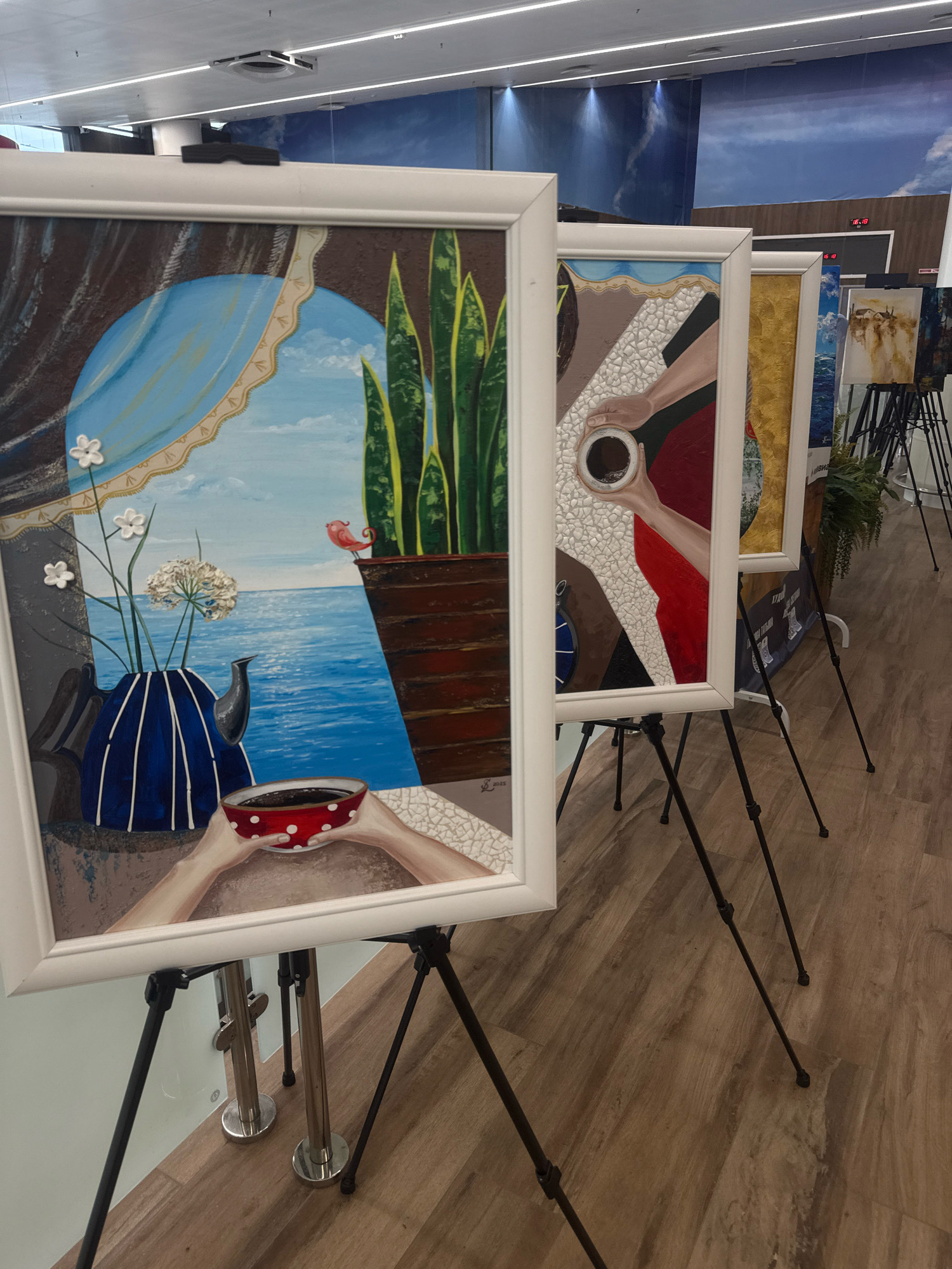 The image shows a row of paintings displayed on easels in what appears to be an indoor exhibition space. The paintings are framed in white. The painting in the foreground depicts a blue vase with white striped patterns, holding white flowers, with a red bird perched on a brown ledge overlooking a blue sea and sky. Below the vase, a pair of hands holds a red bowl with white polka dots filled with a dark liquid. Behind this painting, other artworks are visible, one of which shows a hand holding a cup of coffee. The environment includes a wooden floor and a ceiling with overhead lighting. No people are clearly visible. The provided context states the location is Izhevsk, Russia, however, there are no visual elements in the image that specifically identify this location.