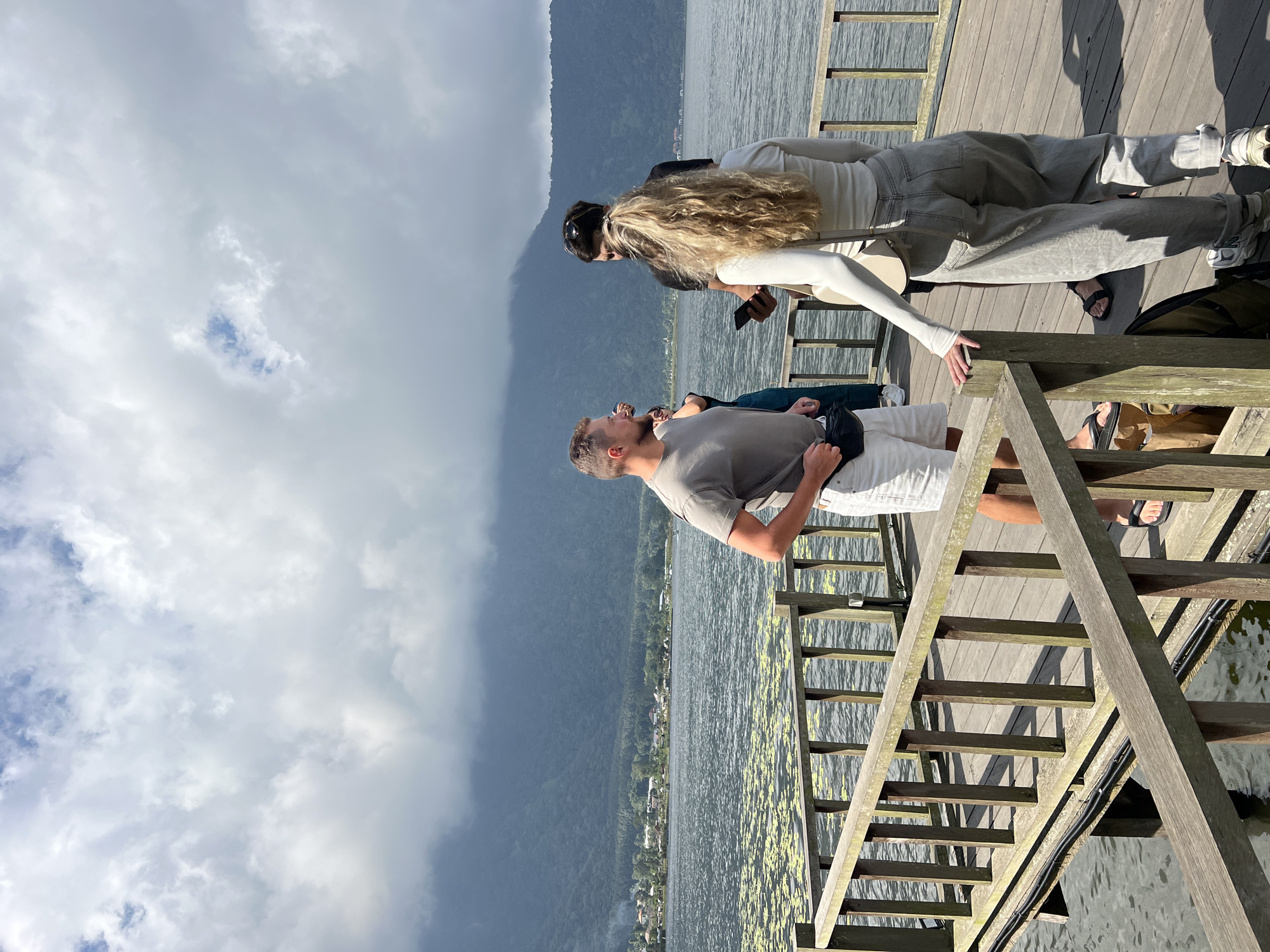 Four individuals are on a wooden pier overlooking a lake with distant mountains under a cloudy sky. A man in a grey t-shirt and white shorts stands on the left, facing left with his hands behind his back. To his right, another person in a dark shirt holds a phone. A woman with blonde hair in a white long-sleeved top and grey pants stands facing away from the camera, holding a phone in her right hand. In the background, a calm lake is visible with patches of green vegetation on its surface, and a treeline with buildings runs along the shore. A large, dark, mist-covered mountain range dominates the background, with scattered white clouds overhead.
