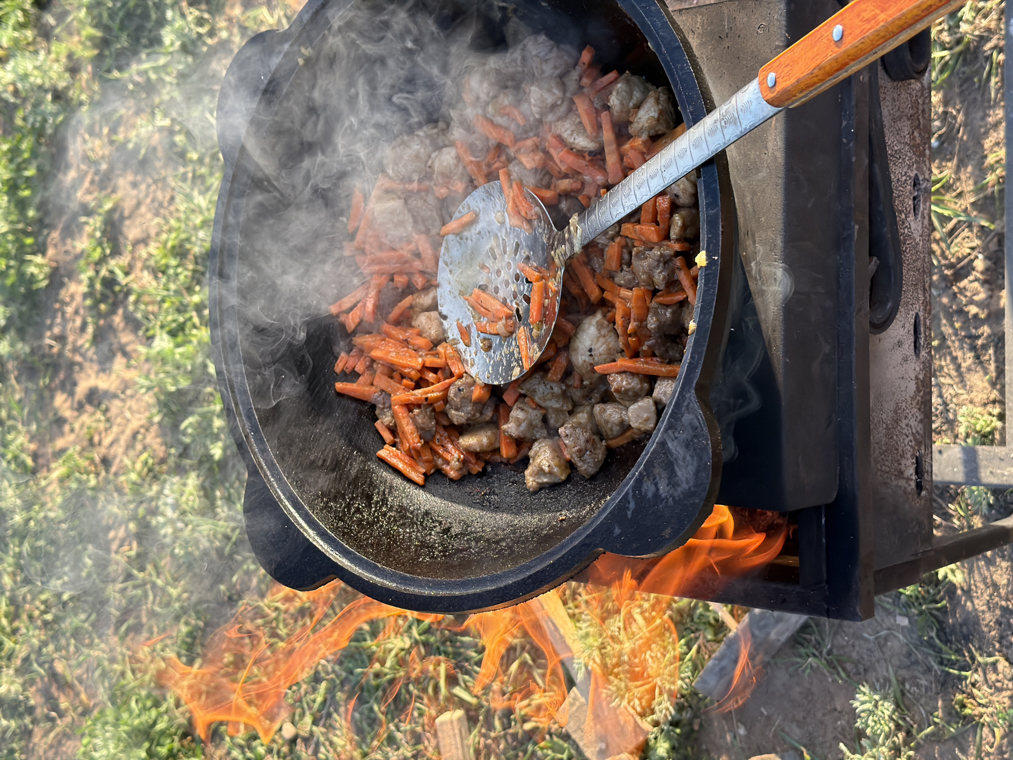A high-angle shot shows food being cooked in a large, black, cauldron-like pot over an open fire. The food inside the pot appears to be a mixture of sliced carrots and pieces of meat, with steam rising from the hot contents. A large, slotted spoon with a wooden handle is inside the pot, presumably used for stirring the food. The pot is positioned on a black, metal stove or cooker, with flames visible underneath, indicating the heat source. The surrounding environment consists of grass, suggesting an outdoor setting. The location of this image is not provided, therefore it is not possible to say with certainty if it is in the city of Chaikovsky, Russia.