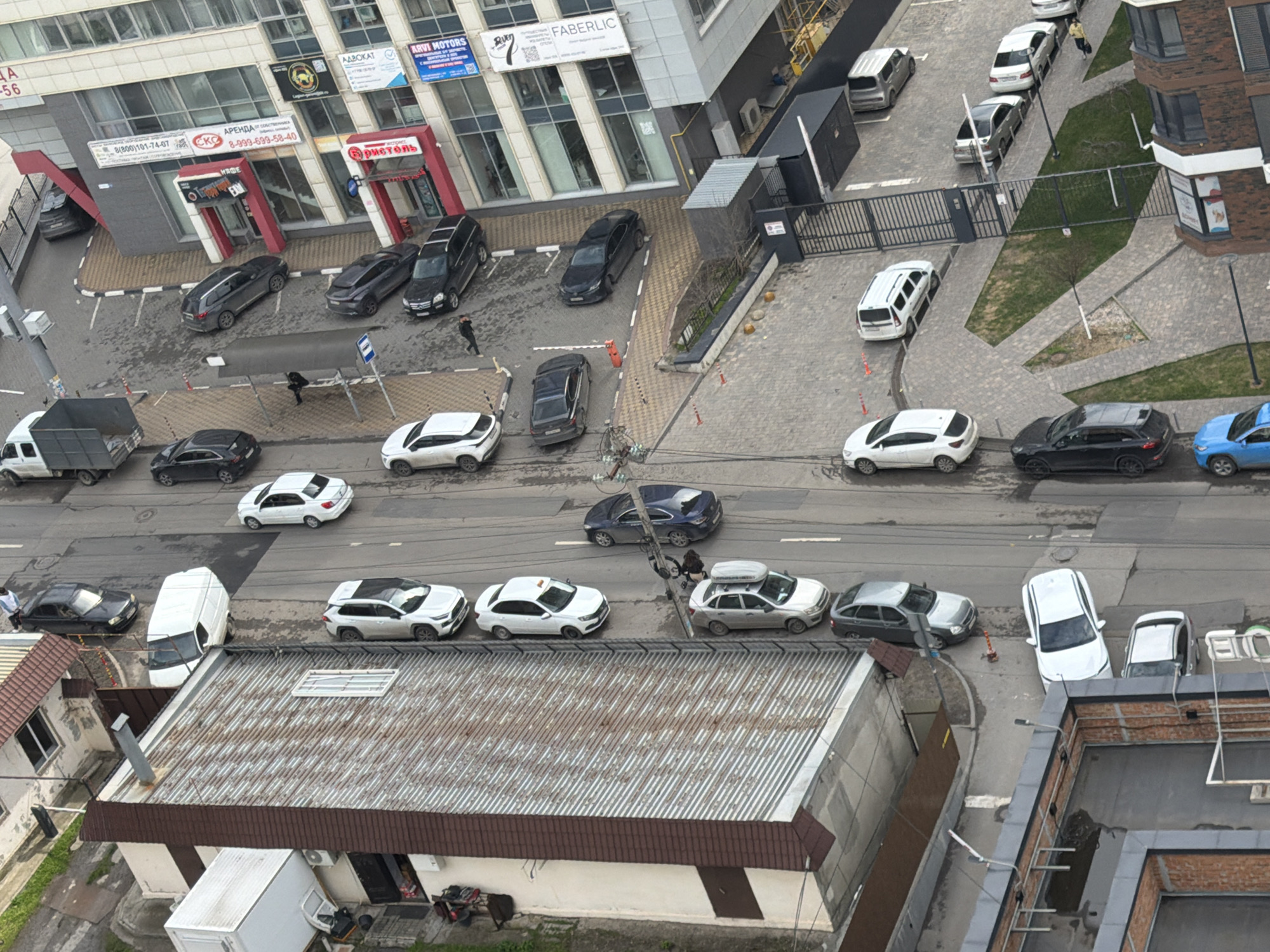The image depicts an aerial view of a street scene in Azov, Russia. A variety of cars are parked along the street, including sedans, SUVs, and a van. A white delivery truck is parked perpendicular to the street. Pedestrians are visible, including one near a bus stop. Several commercial buildings with various signs line one side of the street. The signs indicate businesses such as ARVI Motors, Faberlic, and Бристоль. There are also signs related to property rental and legal services. A low, one-story building with a rusty metal roof sits adjacent to the street. The environment appears to be an urban or suburban setting with a mix of commercial and residential elements.